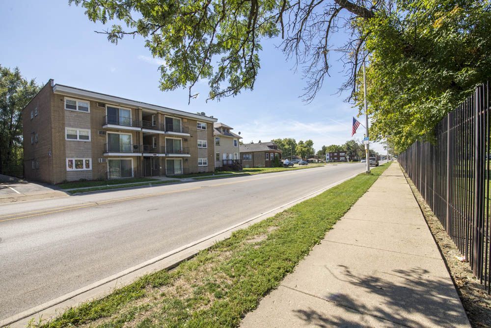 Apartment building next to a road and sidewalk, with a fence and flag.