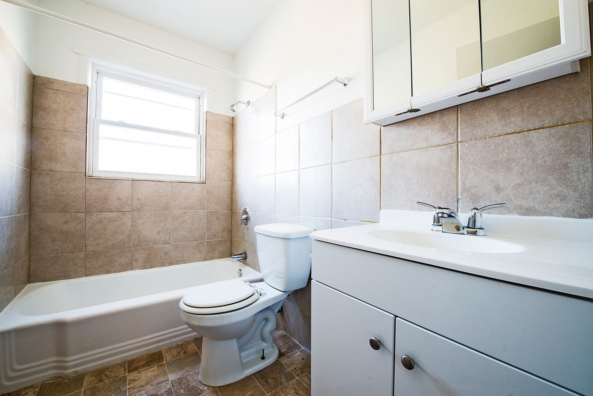 Bathroom with tub, toilet, and vanity. Beige tile walls, window, white fixtures.