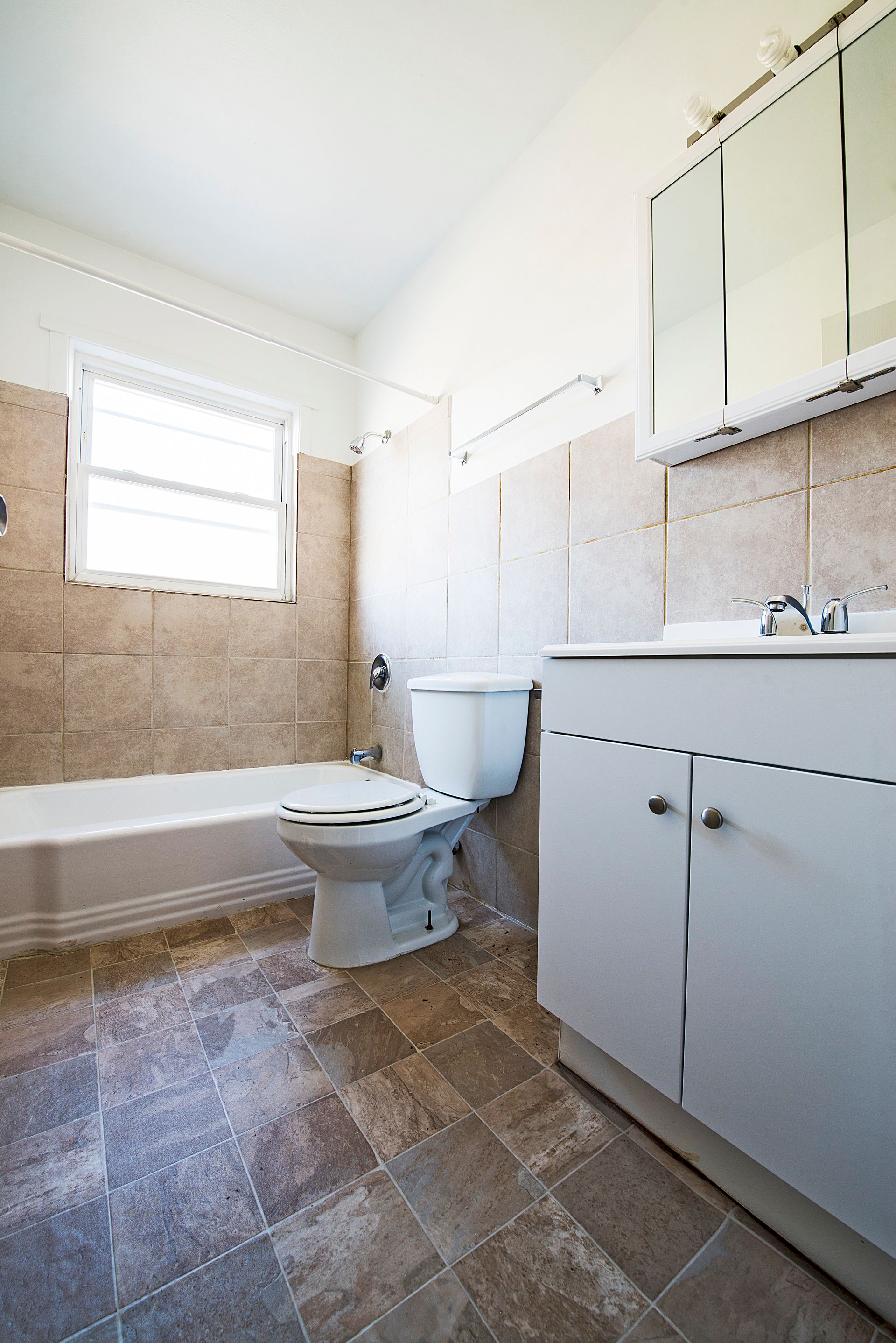 Bathroom with white toilet, vanity, and bathtub, beige tiled walls, and brown tiled floor.