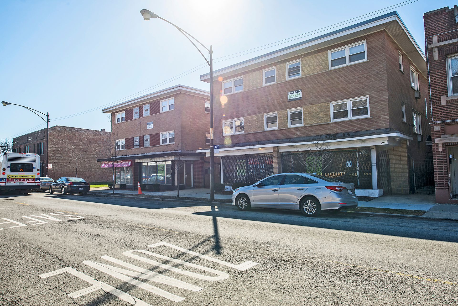 Buildings on a city street. Brick apartment buildings, storefronts with large windows, and parked cars are visible.