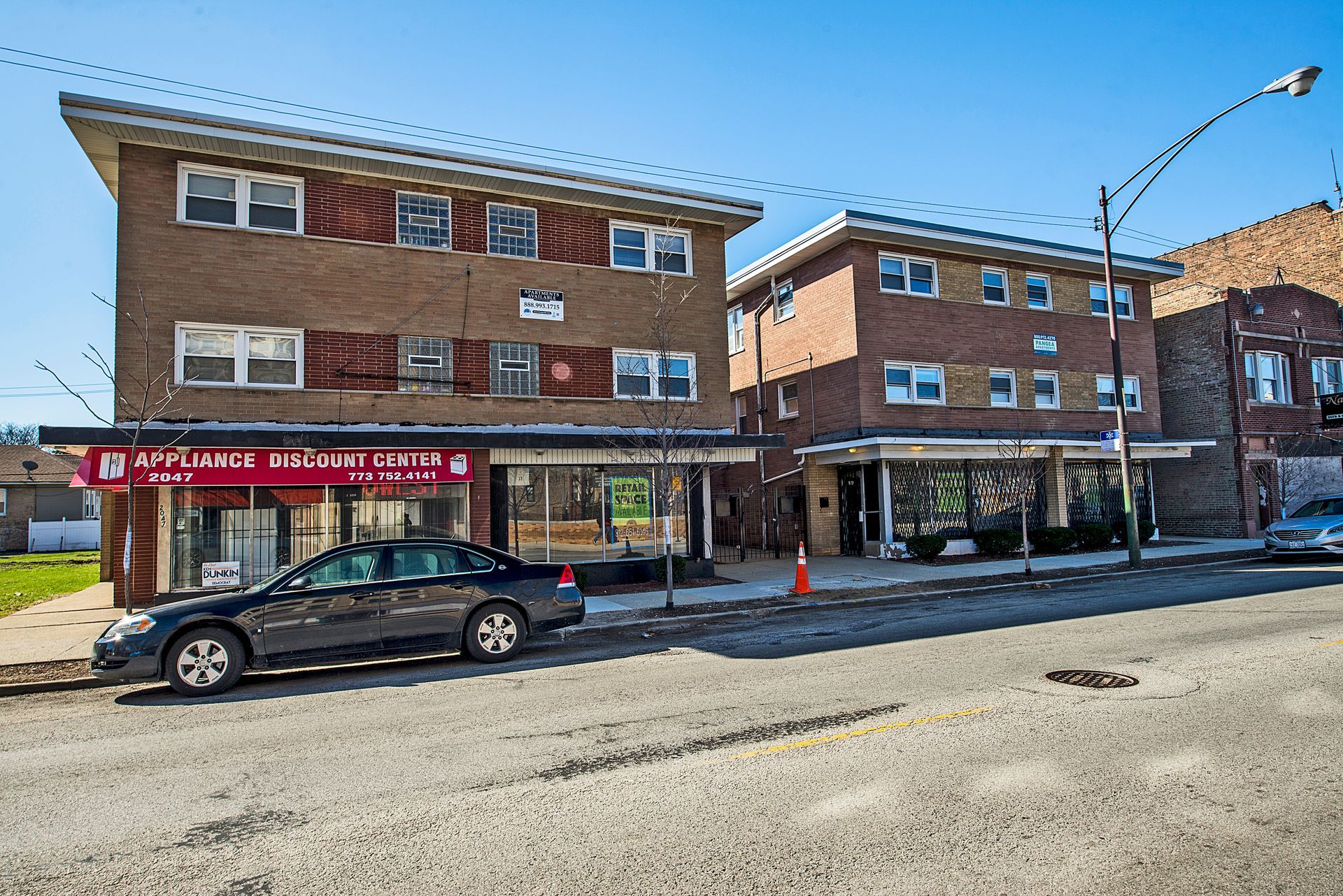 Two-story brick buildings with storefronts on a city street. A parked car sits in front of one.