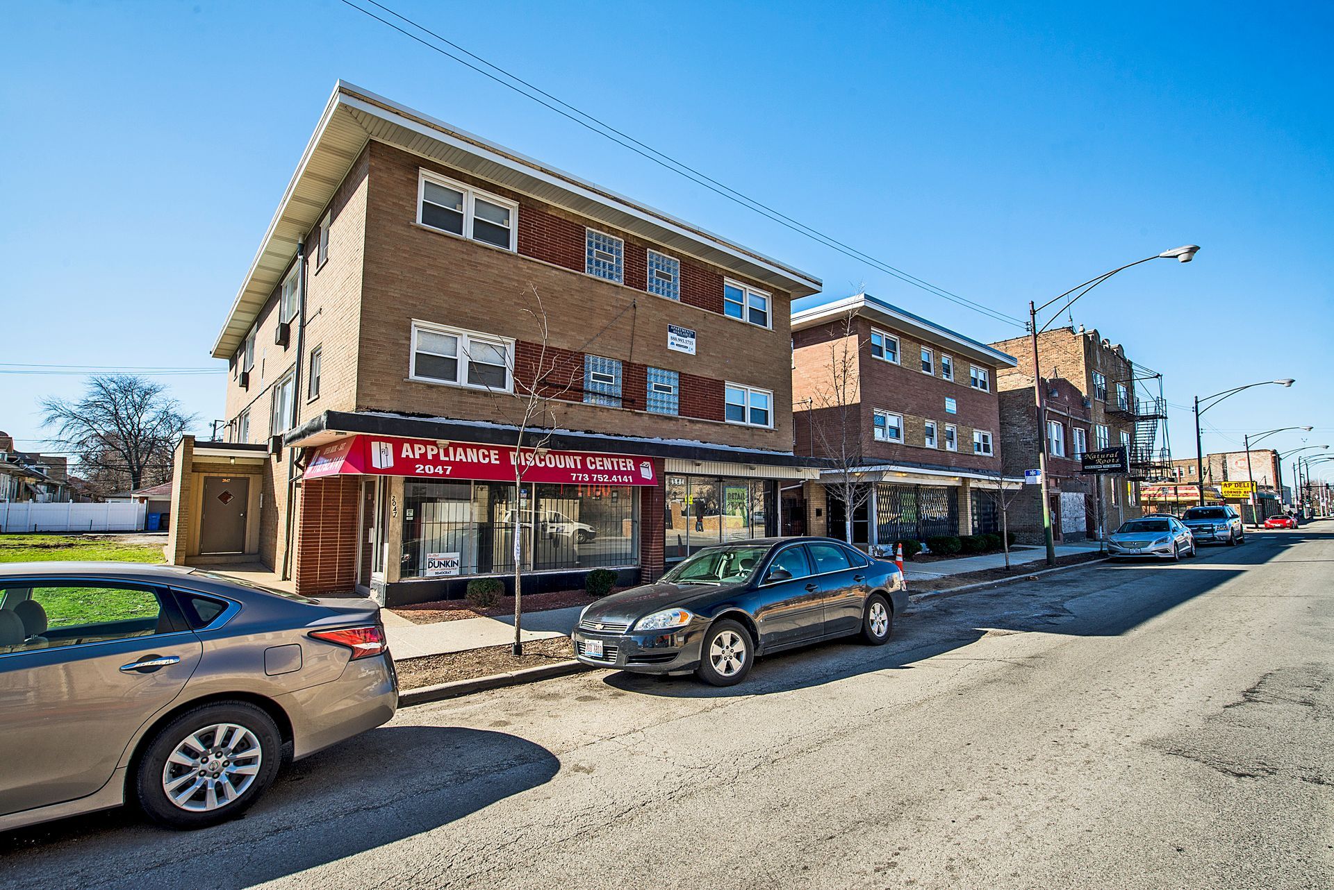 Brick building with businesses on ground level and apartments above, cars parked on street.