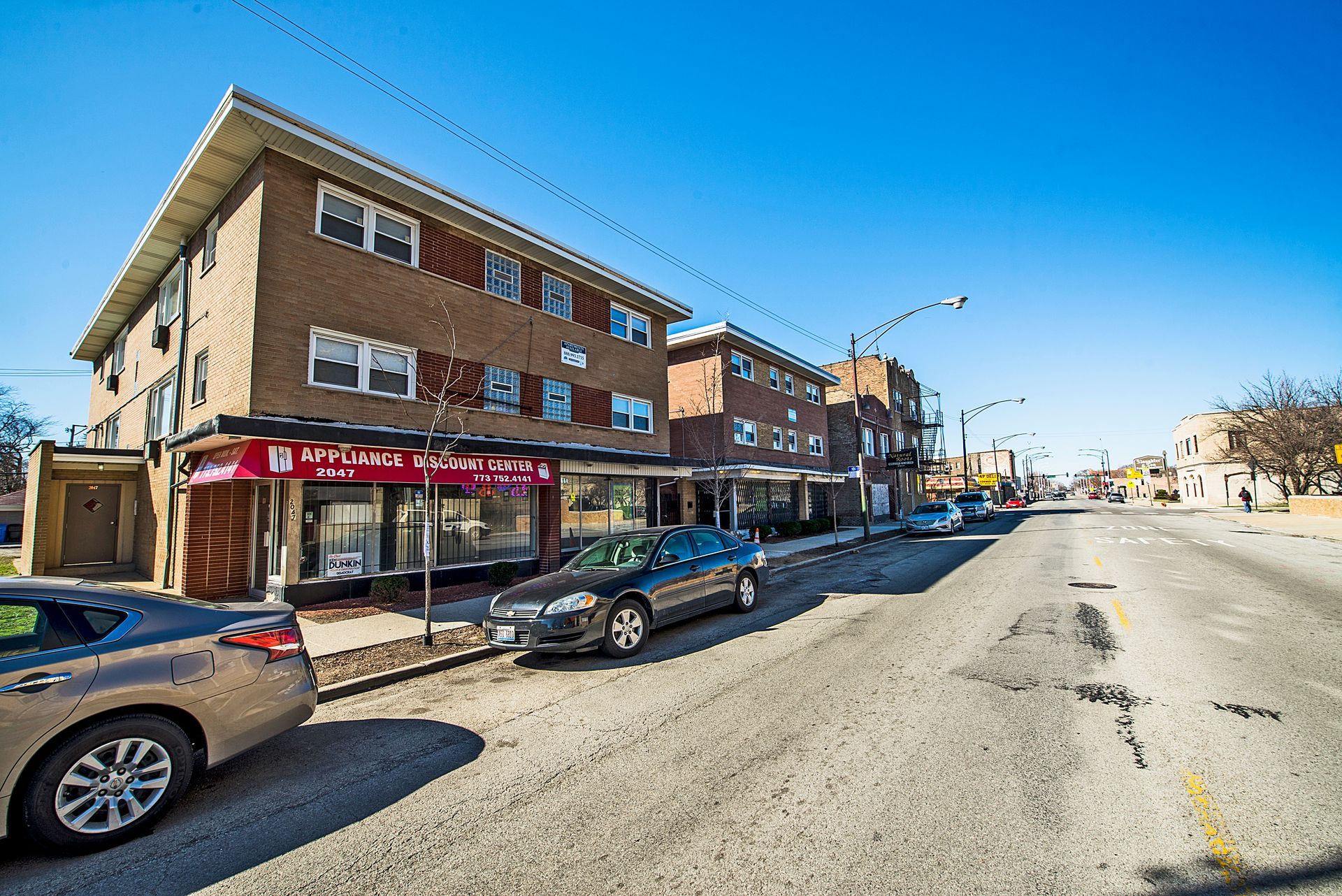 Street view of a brick building with a shop, cars parked, and a cracked road under a blue sky.