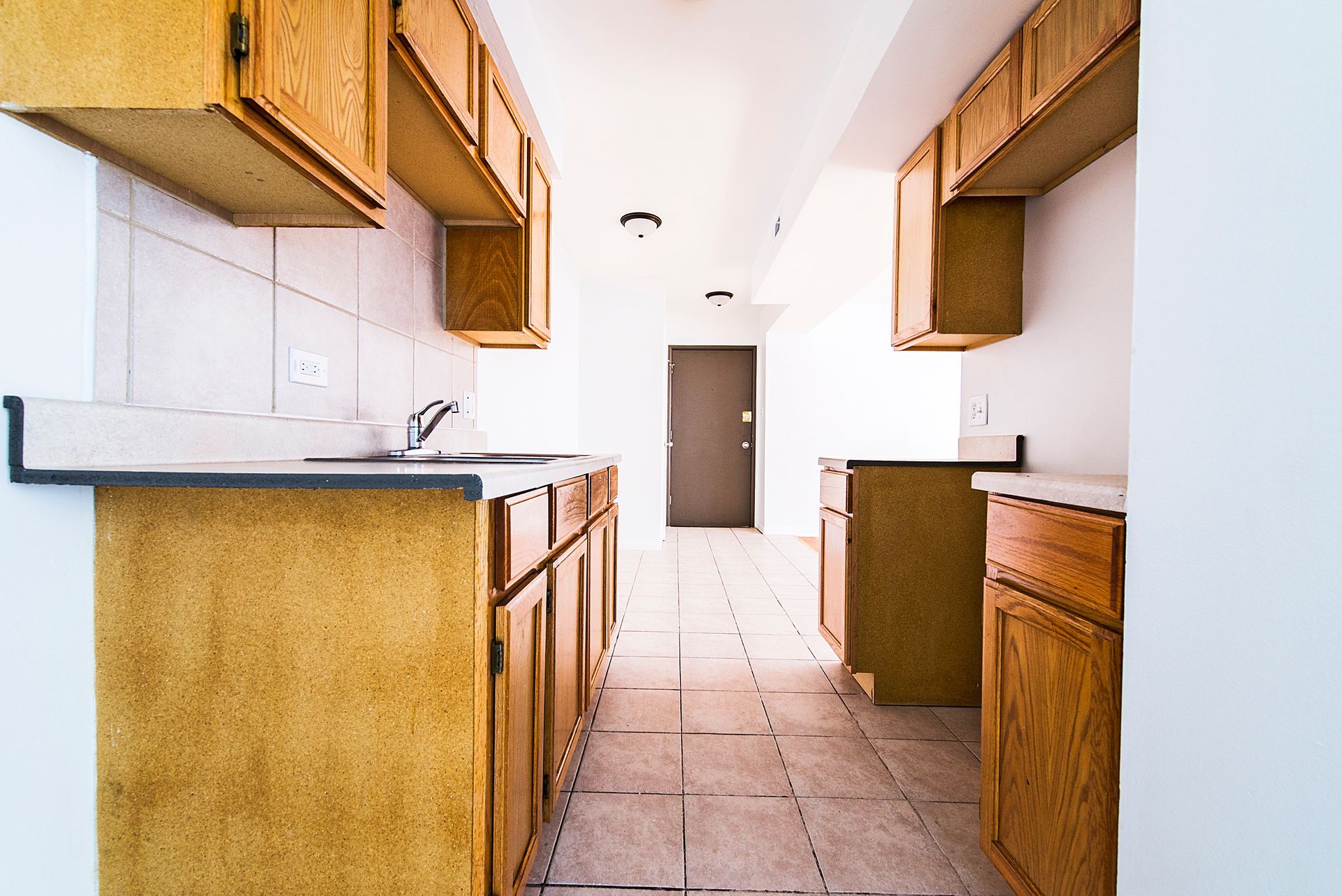 Empty kitchen with light-colored cabinets, countertops, and tiled floor; door at the end of the room.