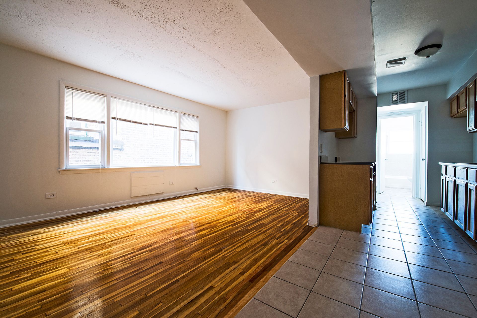 Empty interior with hardwood floor and tile, window with blinds. Kitchen with cabinets and door.