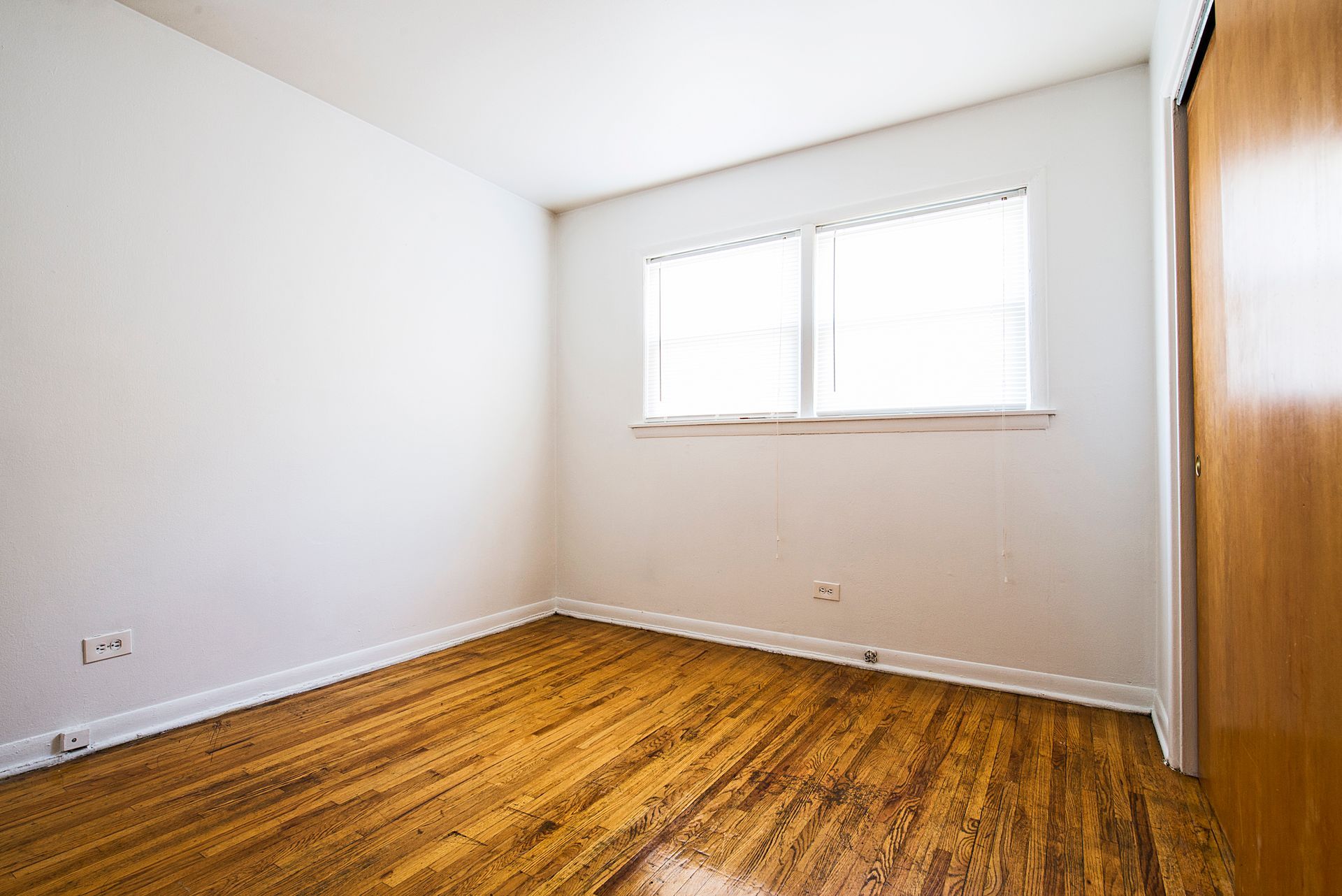Empty room with wood floors, white walls, small window, and a closed wooden door.