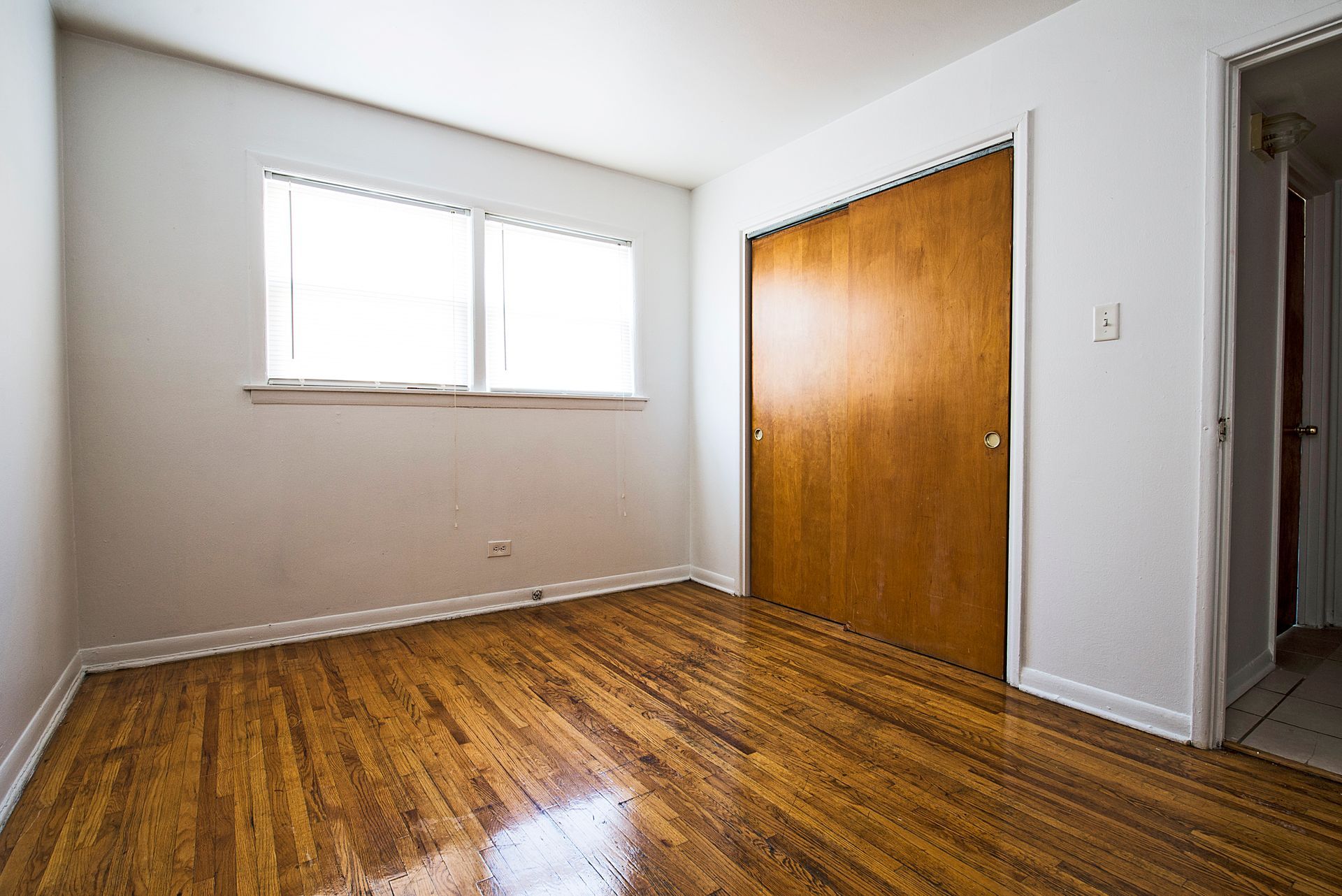 Empty bedroom with wood floors, closet, window, and white walls.