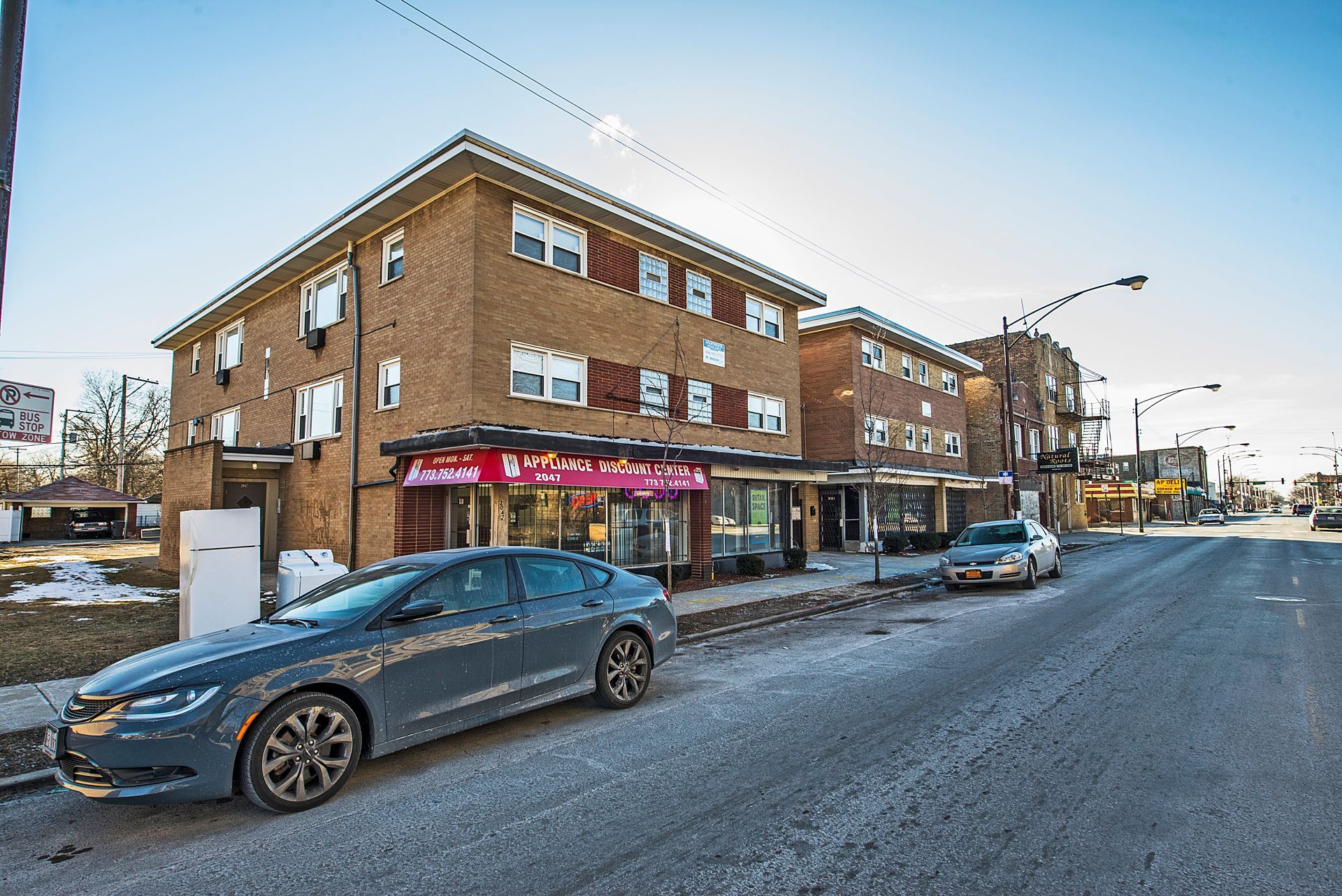 Apartment building with shops on ground floor, gray car parked on street. Sunny day.