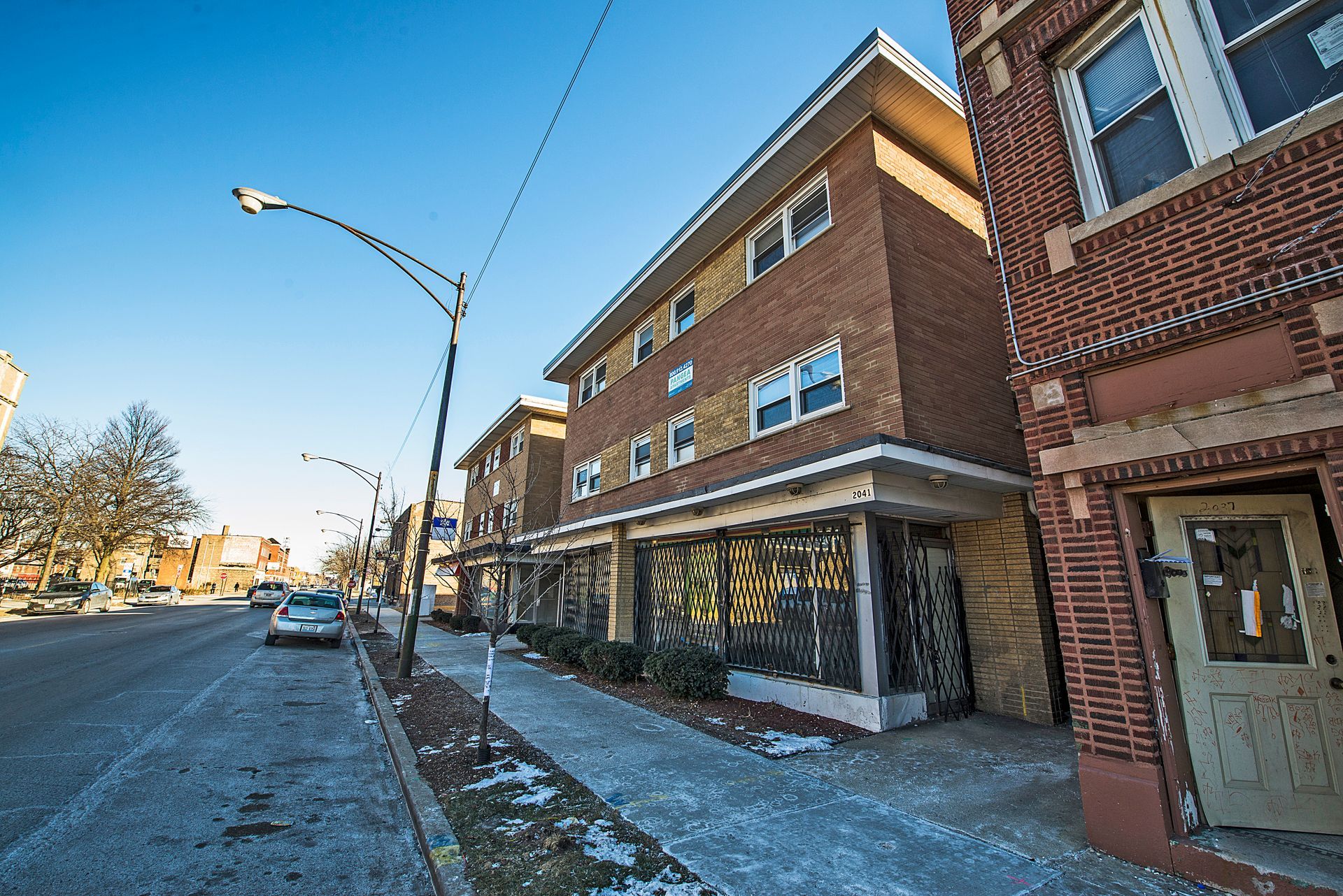 A brick building on a city street with metal bars on the ground level windows and a snow-covered sidewalk.