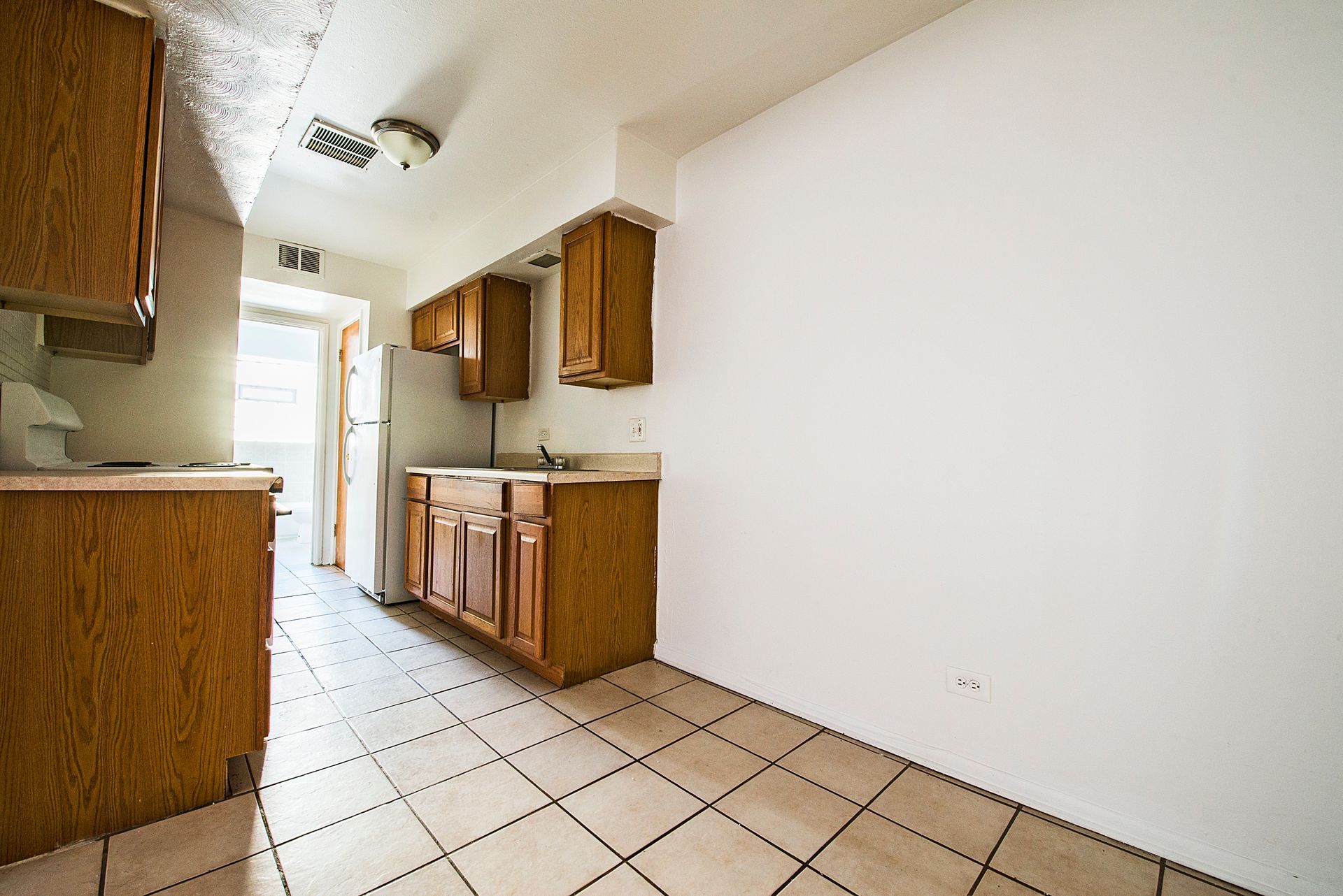 Kitchen with wood cabinets, white walls, and tiled floor. Refrigerator and sink visible.