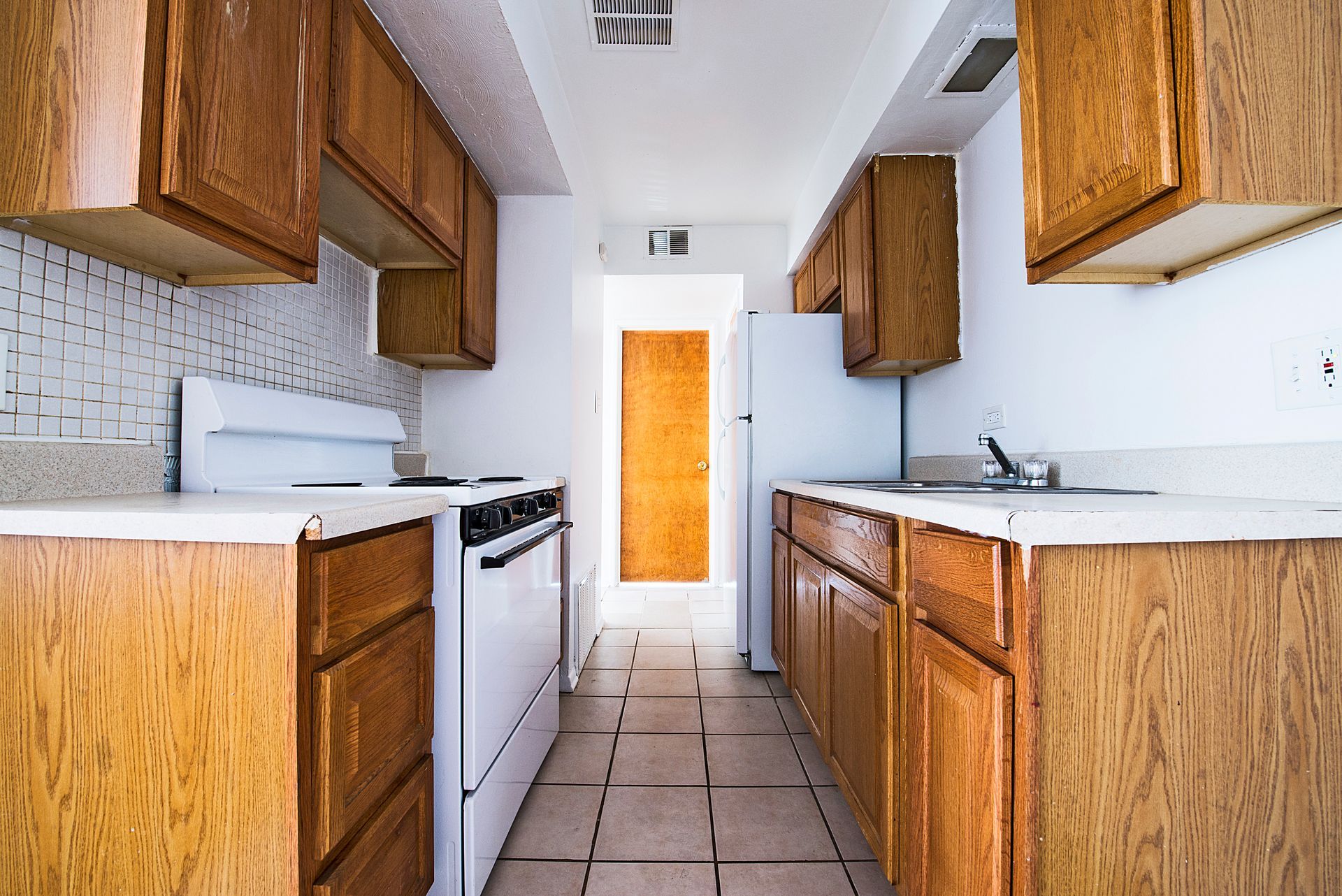 Narrow kitchen with wooden cabinets, white appliances, and a door at the end.