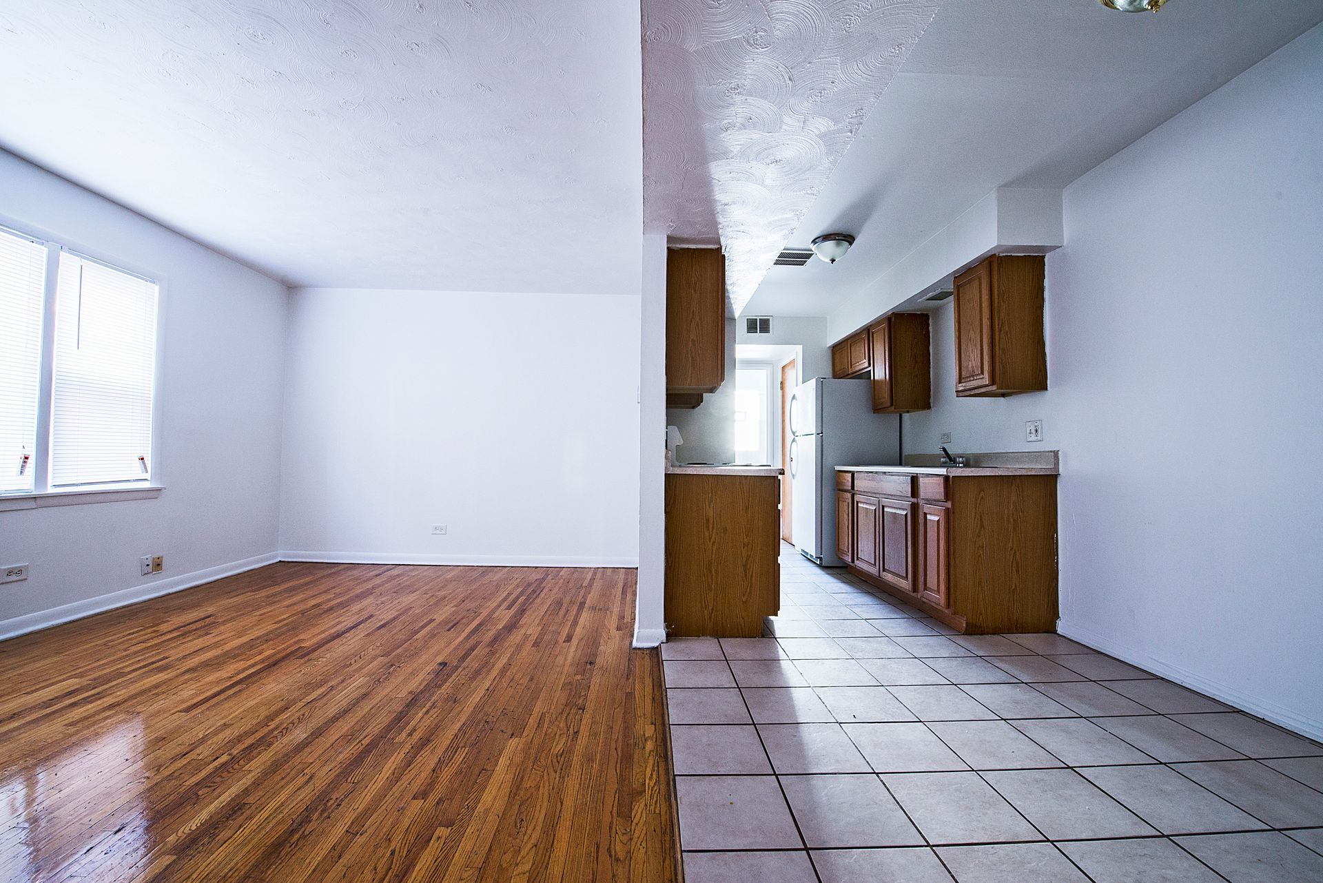 Empty apartment interior: hardwood floor living area, tile floor kitchen with cabinets.