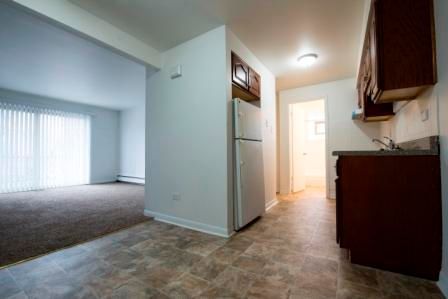 Apartment interior: kitchen with brown cabinets and refrigerator, leading to living room with sliding door.