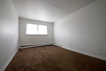 Empty room with white walls, brown carpet, window with blinds.