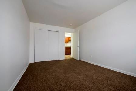Empty room with white walls, brown carpet, and a doorway leading to a kitchen.