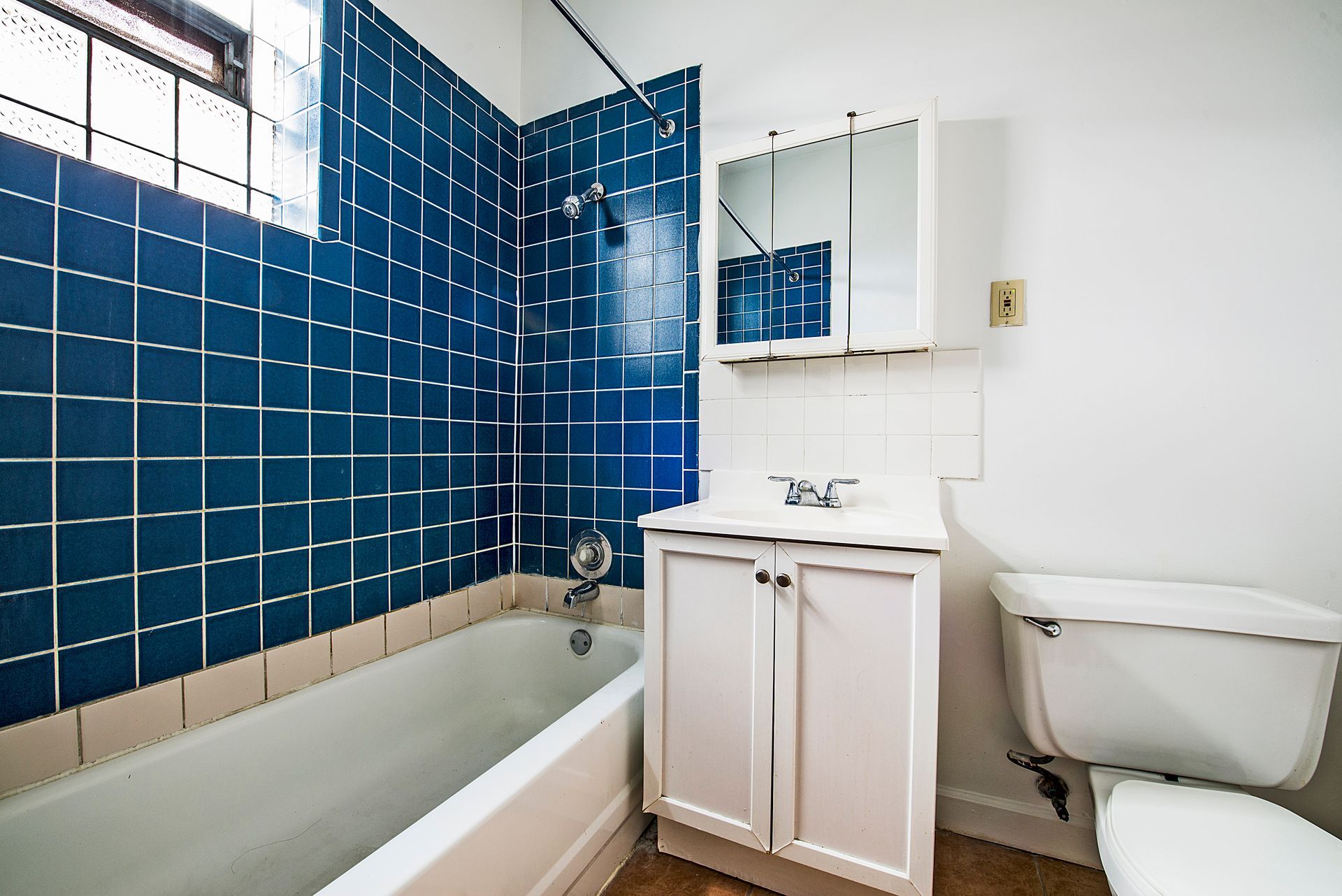 Bathroom with blue tiled shower, white vanity, and toilet. A window is above the tub.