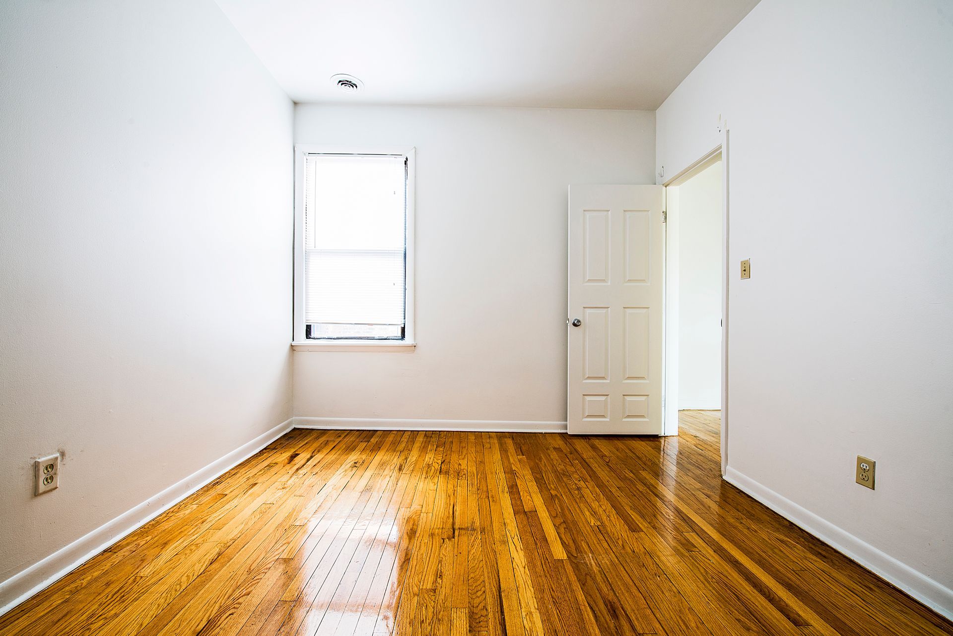 Empty room with wooden floor, white walls, window, and open door.