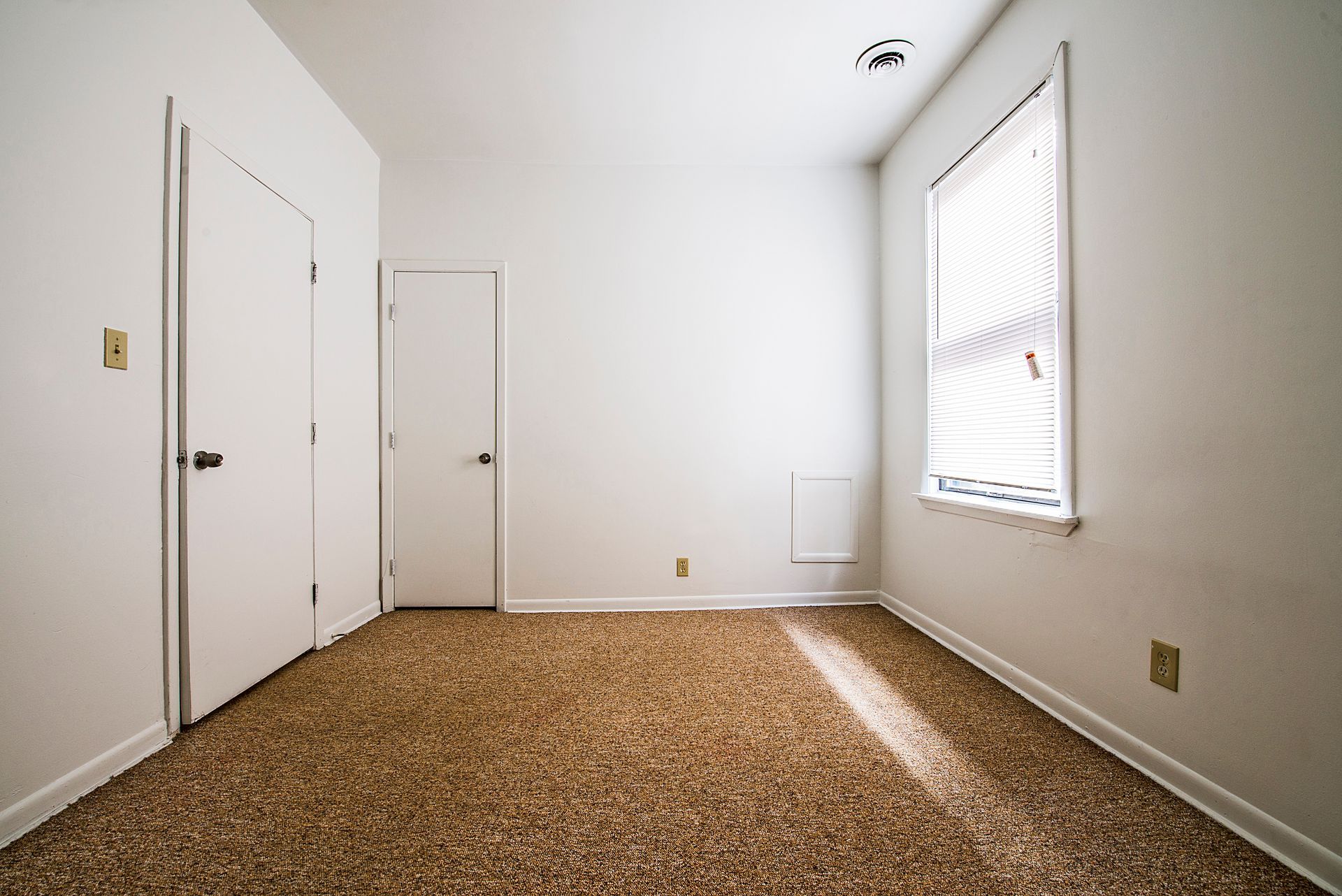 Empty room with white walls, two doors, a window with blinds, and brown speckled carpet.