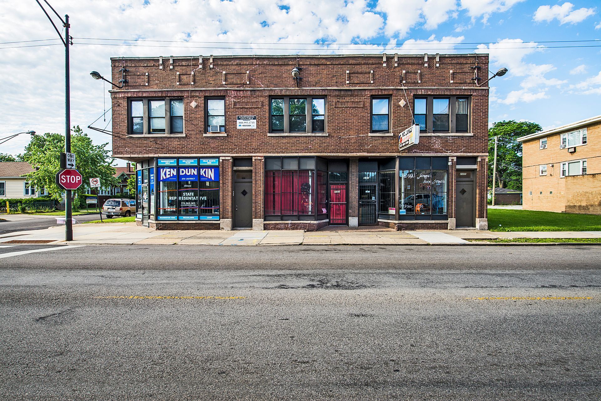 Two-story brick building with storefronts on a city street. Blue sky and residential homes in the background.