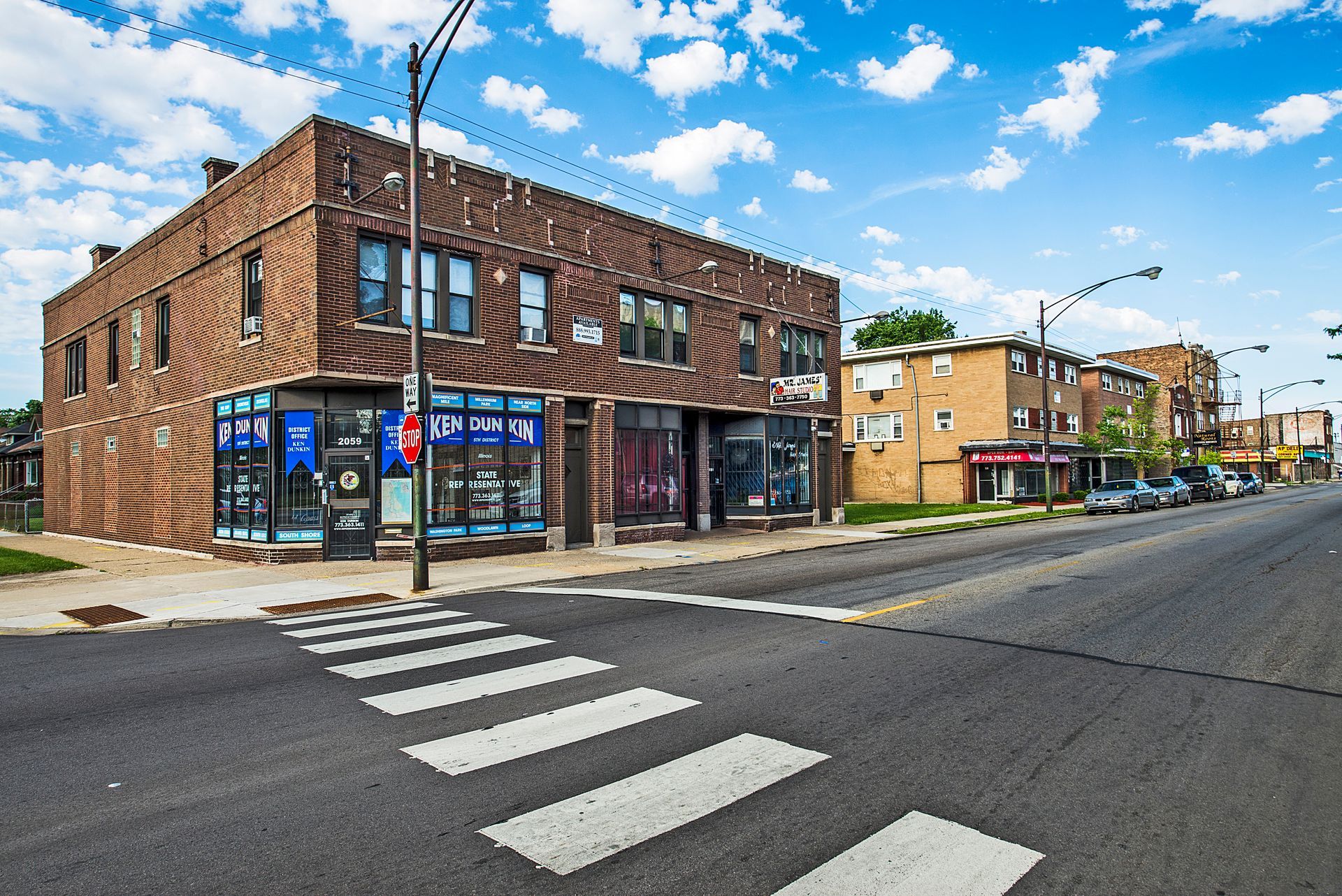 Brick commercial building on a street corner with crosswalk. Blue storefronts below, other buildings line the street.