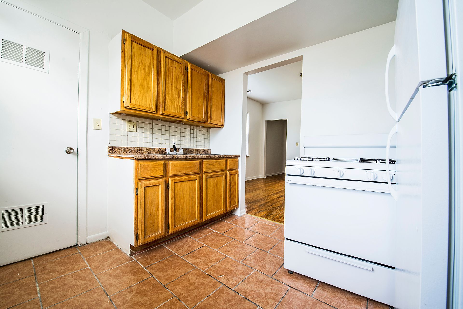 Kitchen with wooden cabinets, white appliances, and a door leading to a hallway with wood flooring.