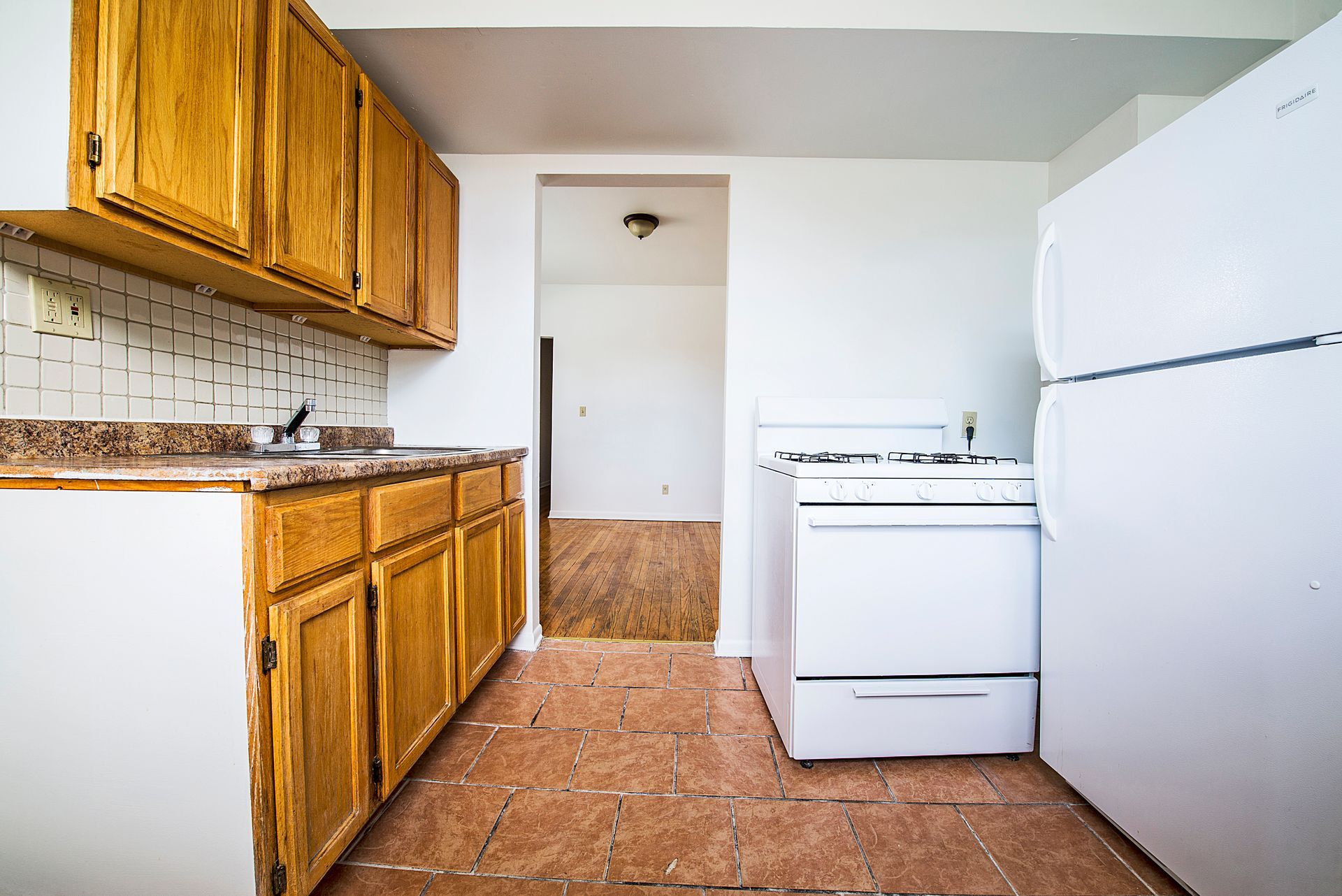 Kitchen with wooden cabinets, white appliances, and a doorway to another room. Brown tile floor.