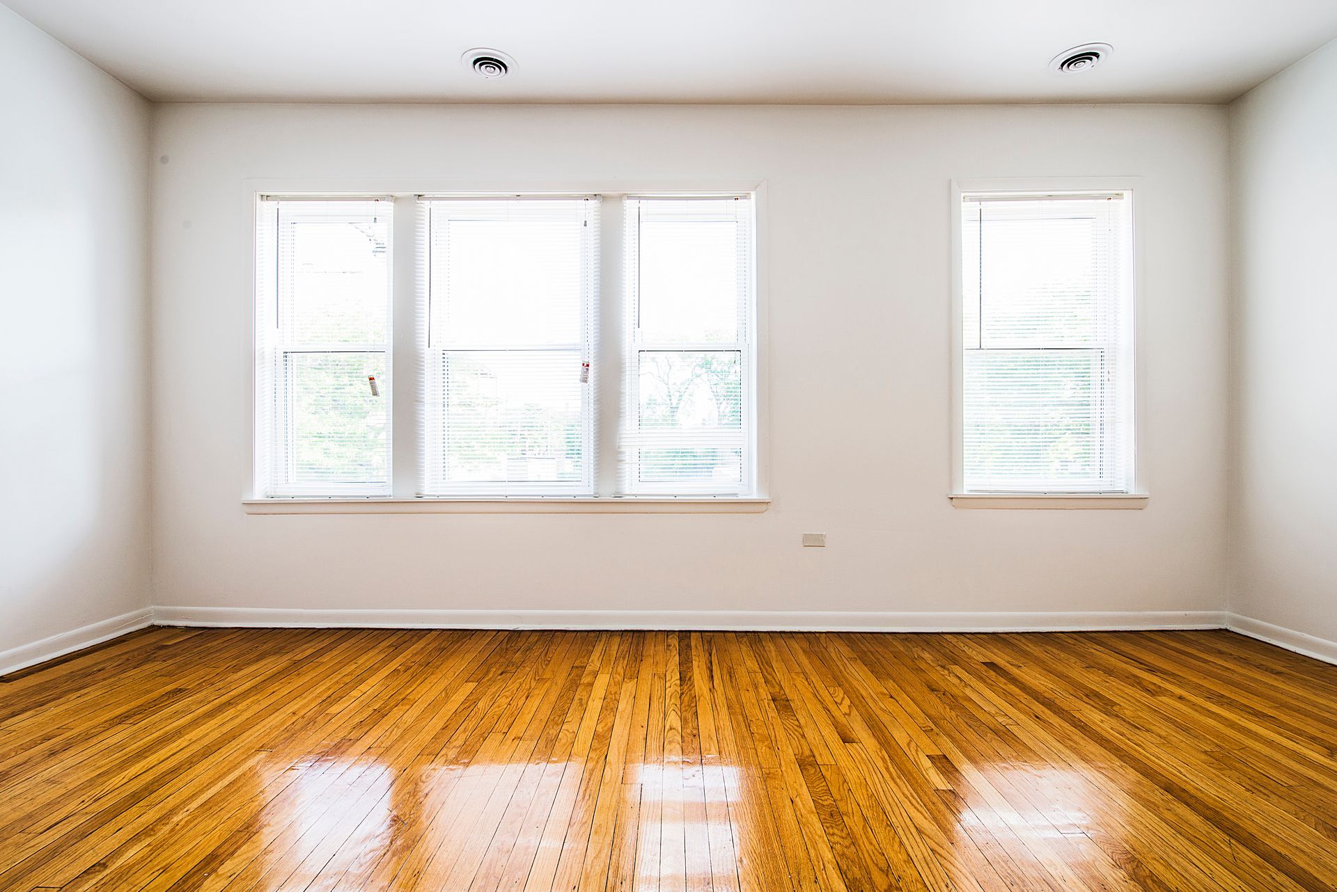 Empty room with hardwood floors, white walls, and windows with blinds.