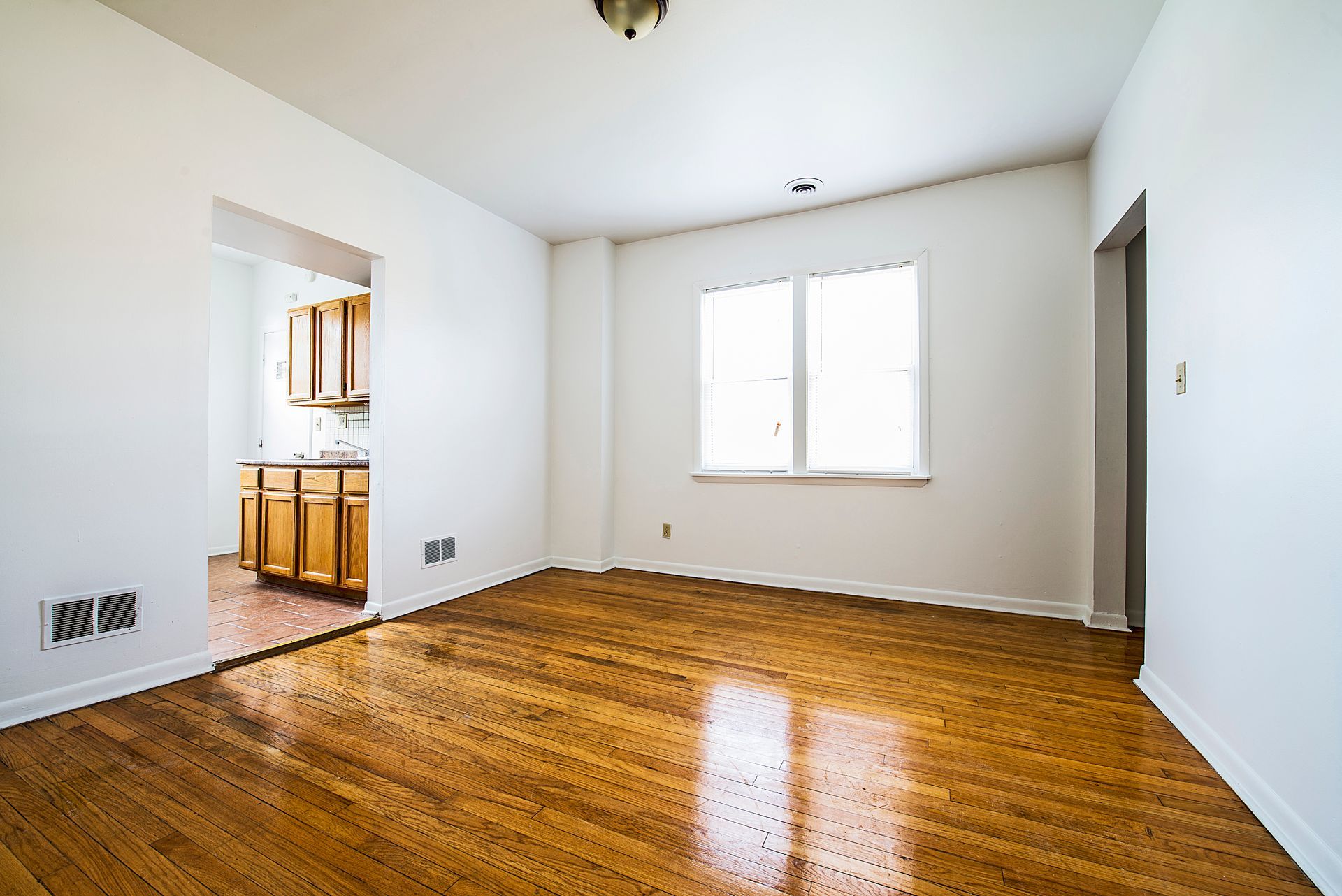 Empty room with hardwood floors, white walls, and a view into a kitchen.