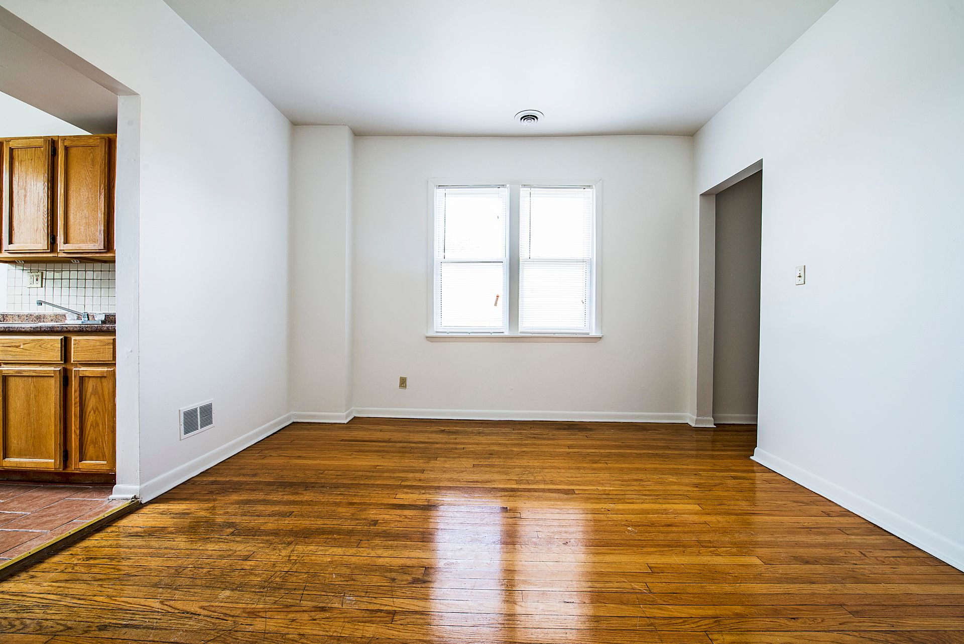 Empty room with hardwood floors, white walls, and a window. Kitchen visible to the left.