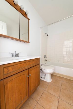 Bathroom with wooden vanity, white toilet, and tile floor. White walls, a mirror, and a shower/tub.