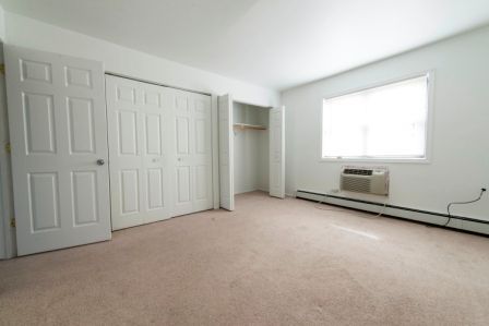 Empty bedroom with white walls, beige carpet, closed closet doors, and a window.