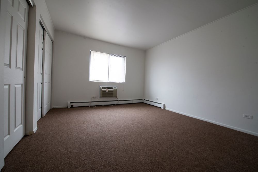 Empty room with brown carpet, white walls, closet doors, and a window with a small air conditioning unit.