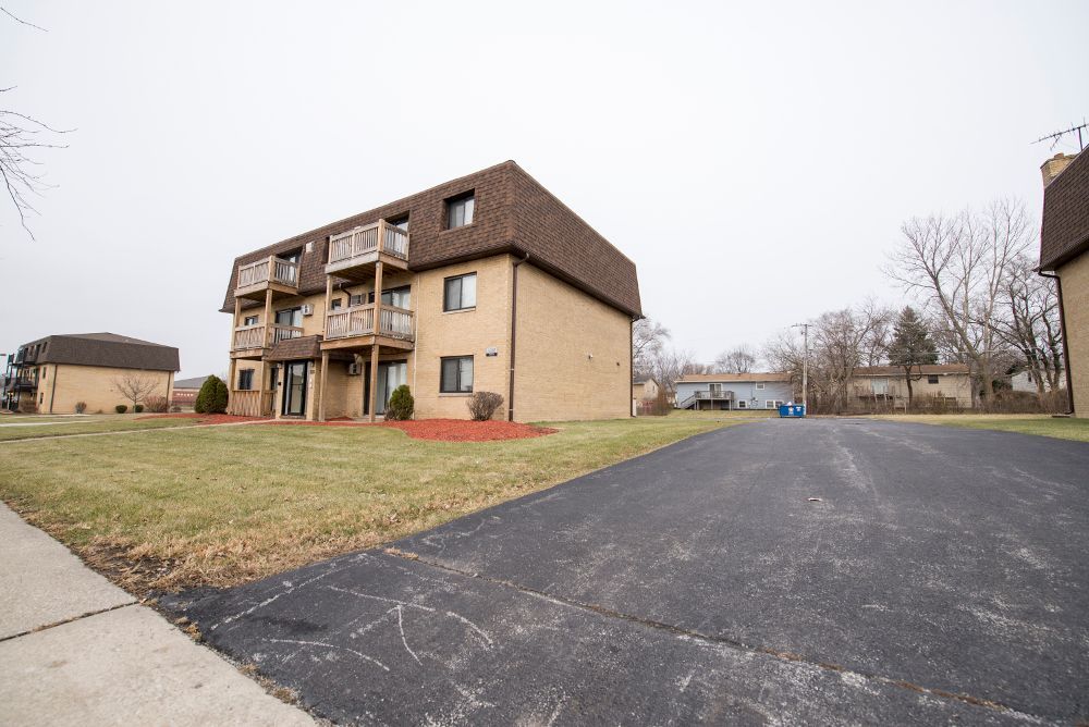 Apartment building with brown roof and balconies, on a cloudy day. Driveway in foreground.