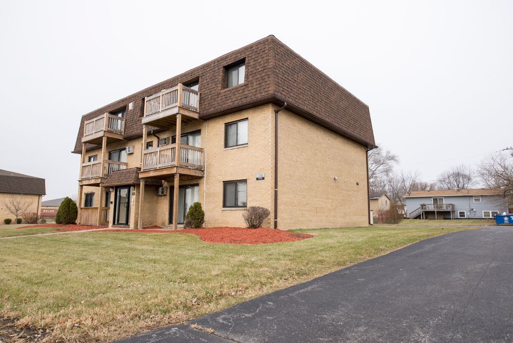 Apartment building with brown roof and balconies, beige brick exterior, on a grassy lot with a paved driveway.