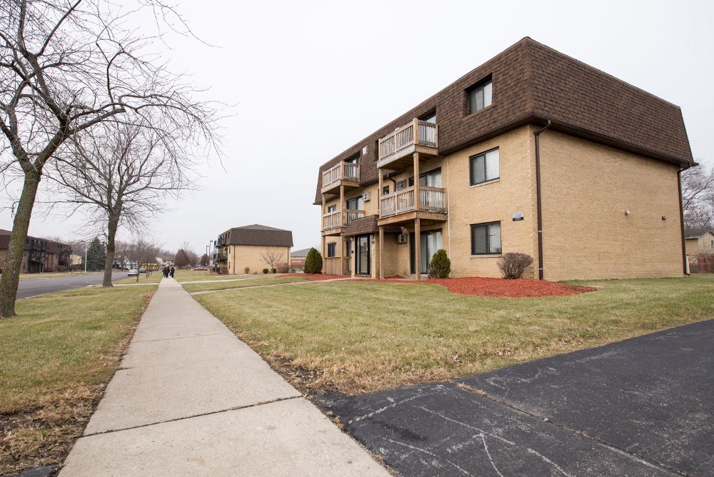 Apartment building with brown roof and balconies, next to a sidewalk and grassy area. Gray sky.