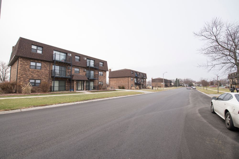 Apartment buildings line a street on an overcast day; a white car parked on the right.