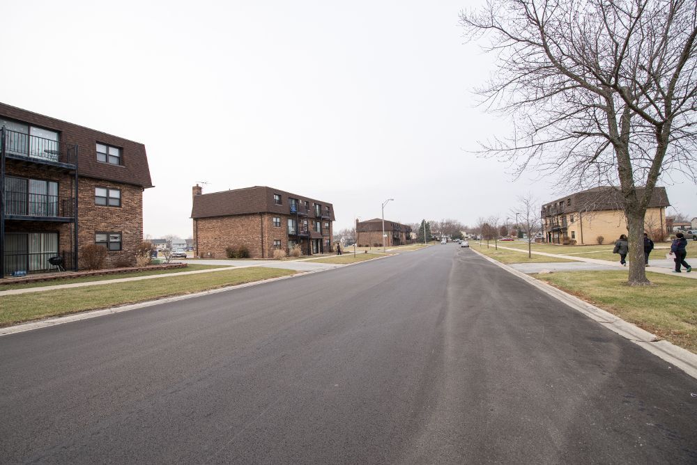 Street lined with brick apartment buildings and bare trees on a cloudy day. Two people walk on the sidewalk.