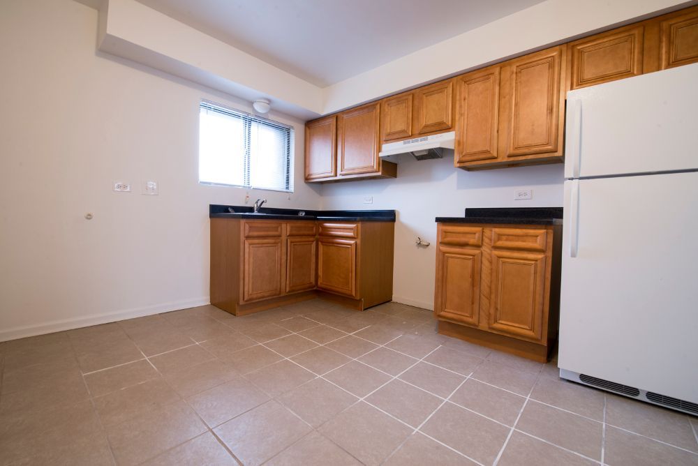 Empty kitchen with wood cabinets, black countertops, and white refrigerator on tile floor.