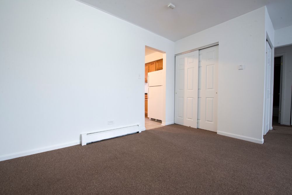 Empty room with white walls, brown carpet, and a closet. Kitchen visible in the background.