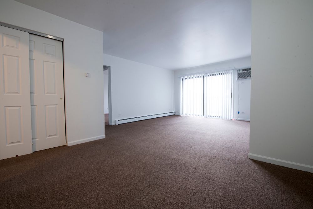 Empty living room with brown carpet, white walls, closet doors, and sliding glass door.