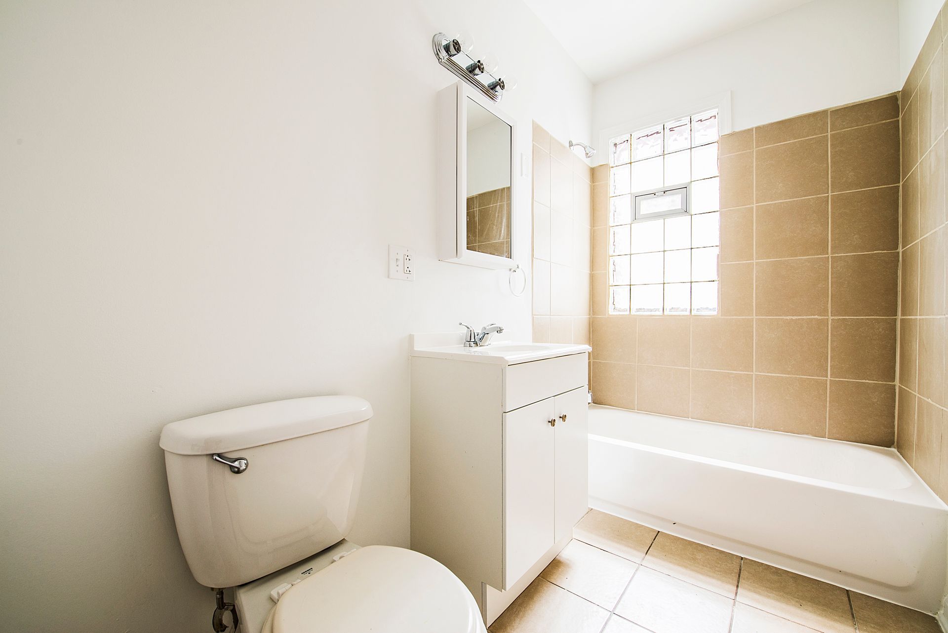 Bathroom with white toilet, vanity, and bathtub; tan tiled walls, and a window.