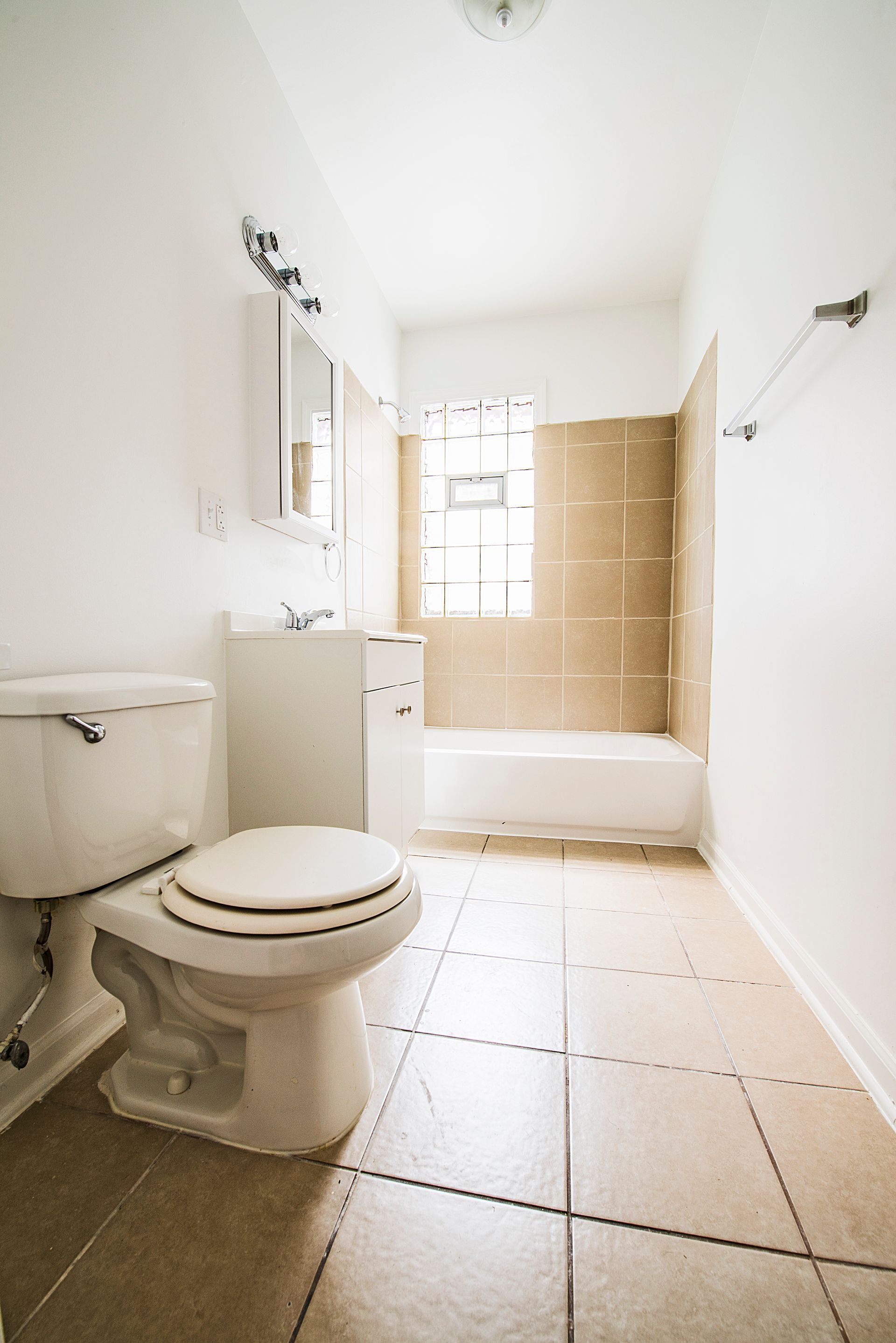Bathroom with white toilet, sink, and bathtub. Beige tiled floor and walls. Natural light from a window.