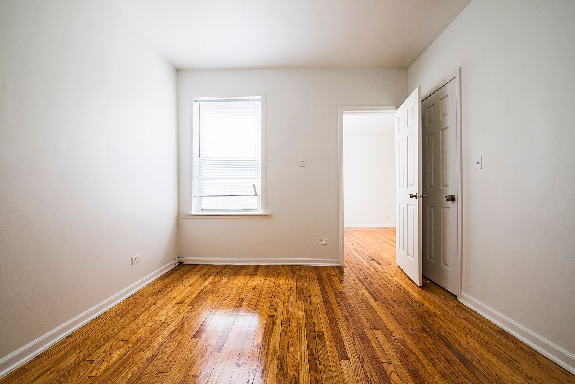 Empty room with hardwood floors, a window with blinds, and an open doorway.
