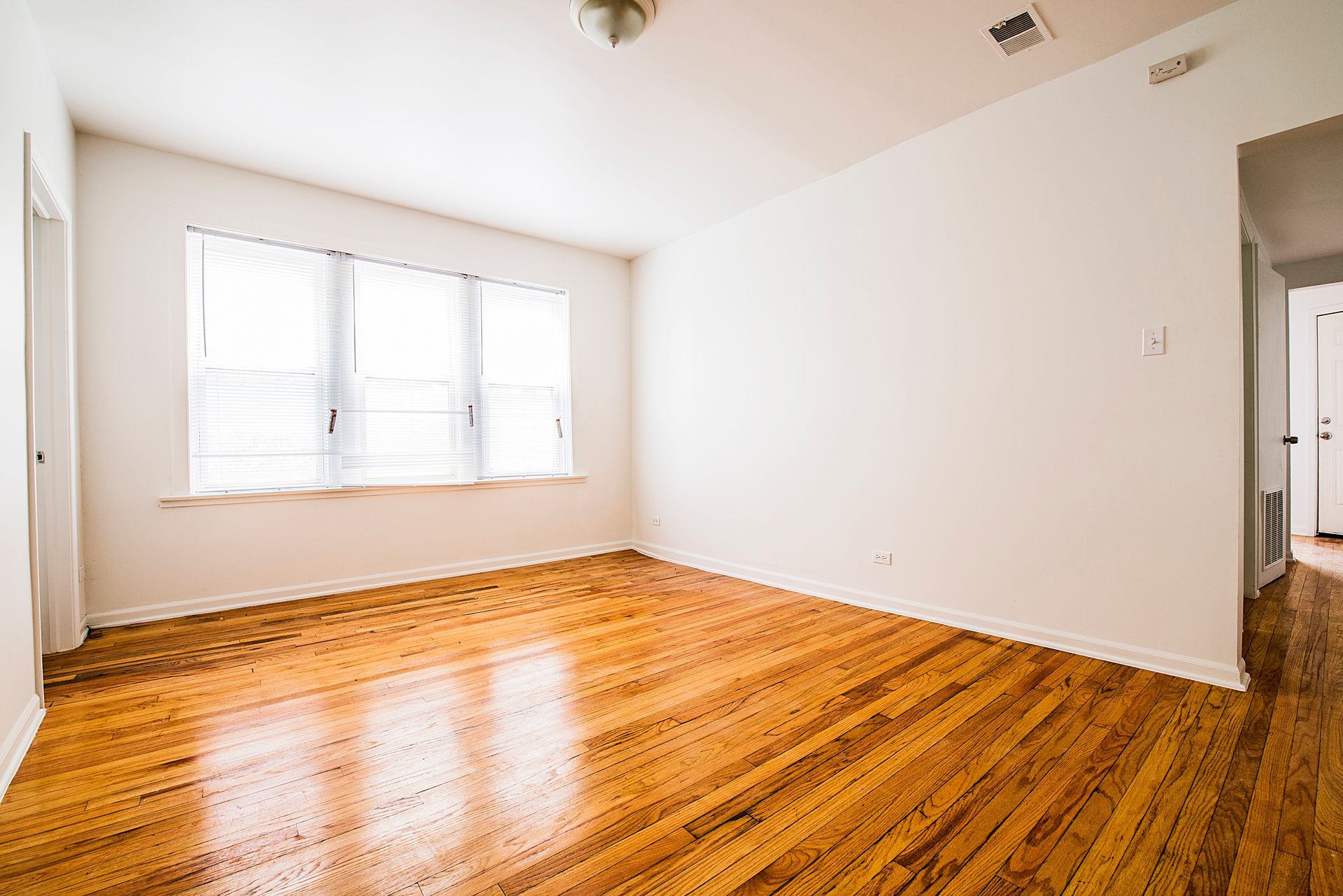 Empty room with hardwood floors, a window with blinds, and white walls.