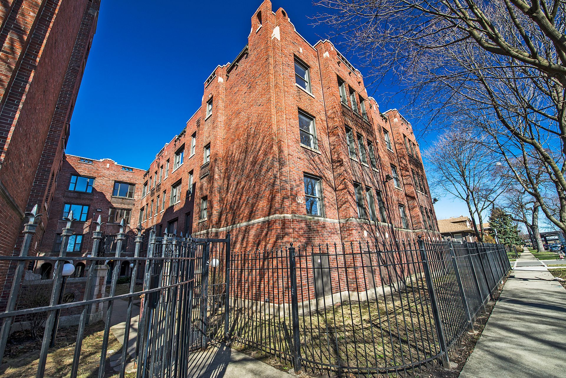 Red brick building covered in ivy behind a black wrought-iron fence on a sunny day.