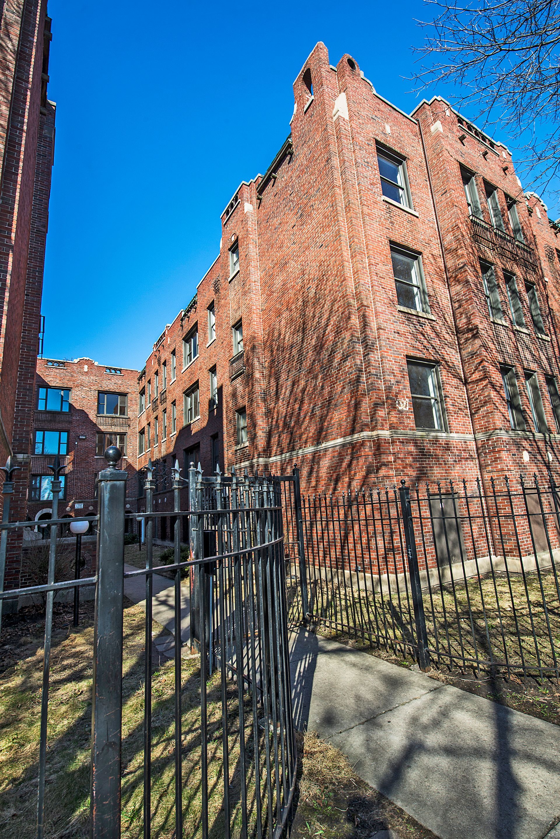 Red brick apartment building with black wrought iron fence against a bright blue sky.