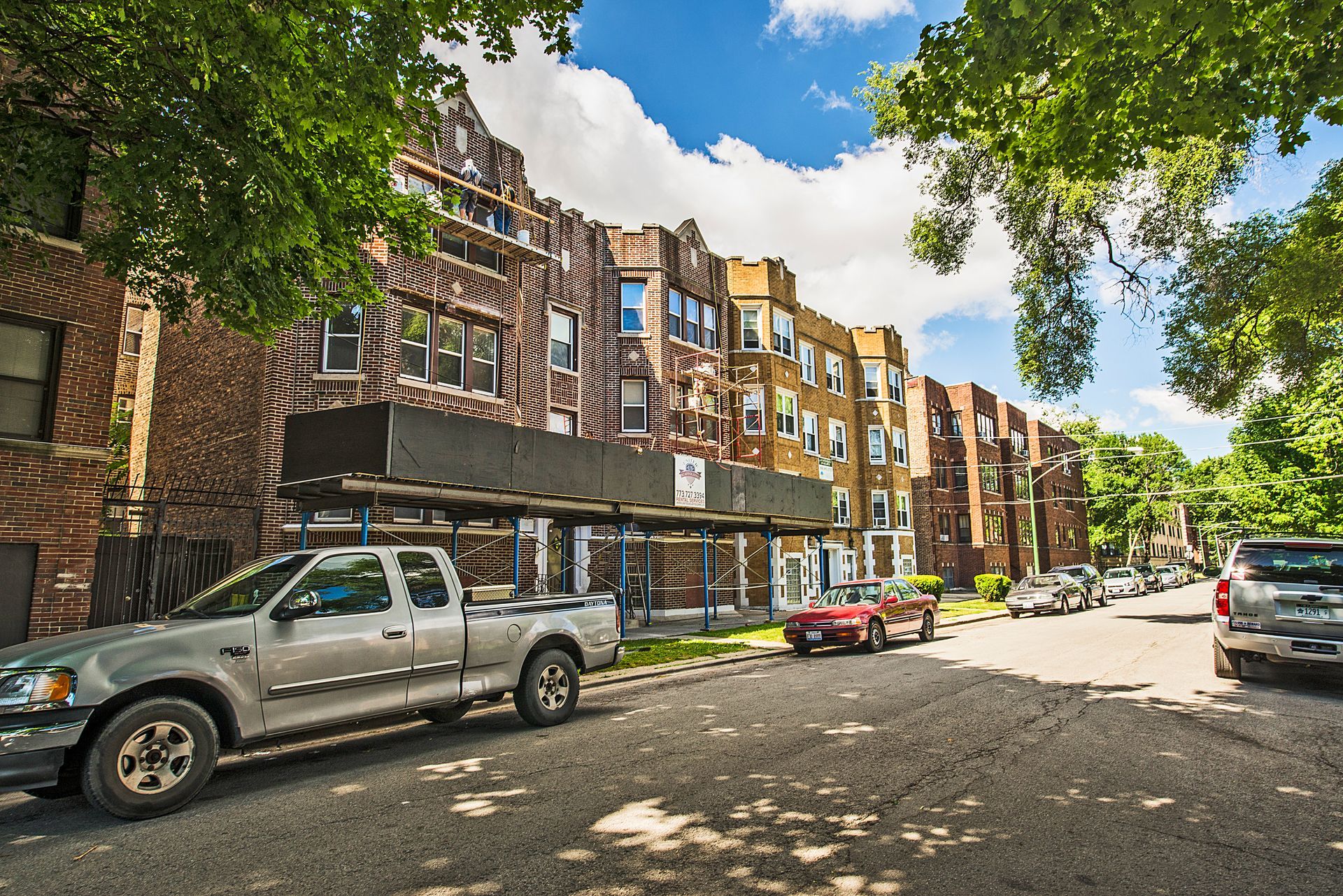 Street view of brick apartment buildings with parked cars and trees. Sunny day.