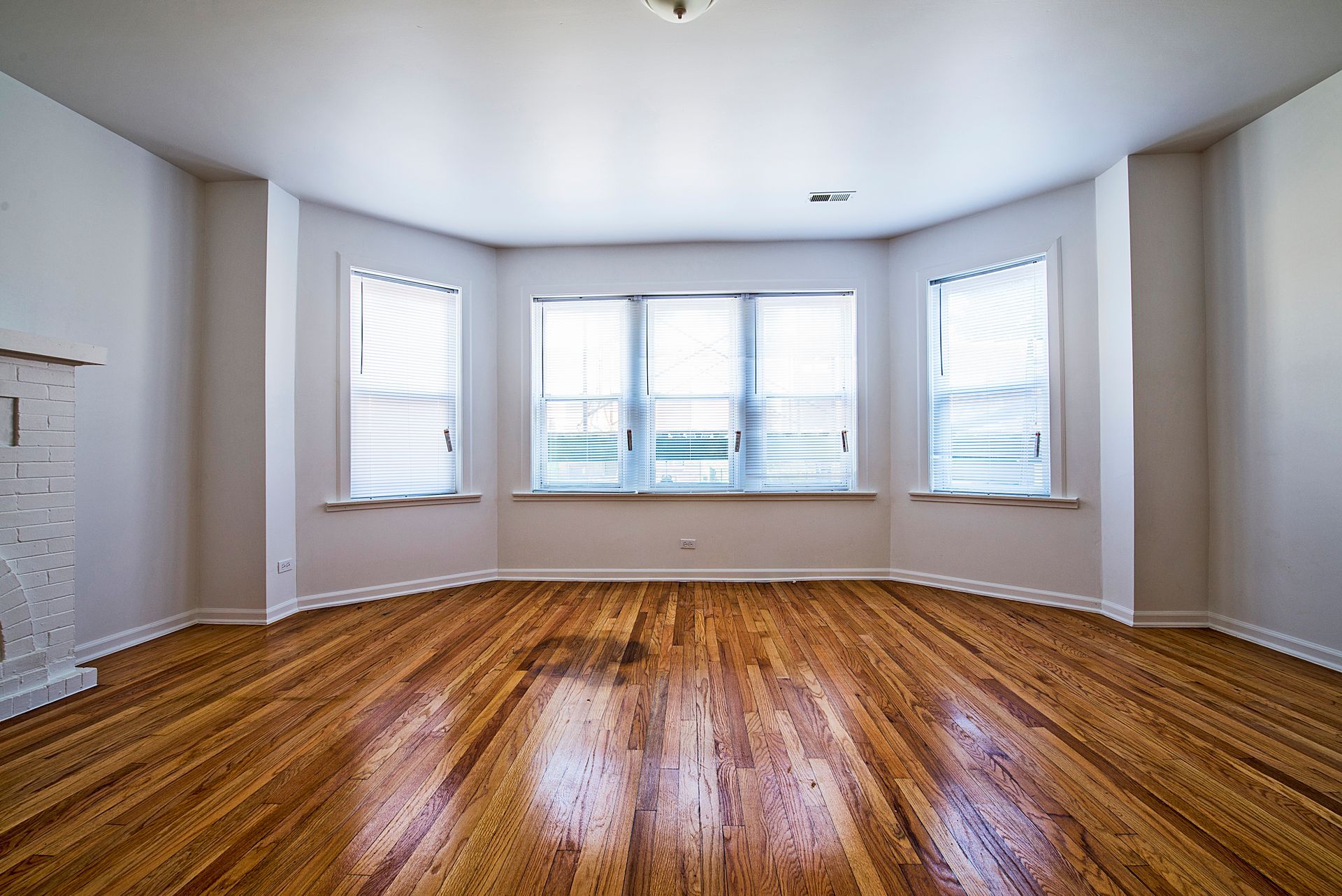Empty room with wooden floor, bay windows with blinds, and white walls.