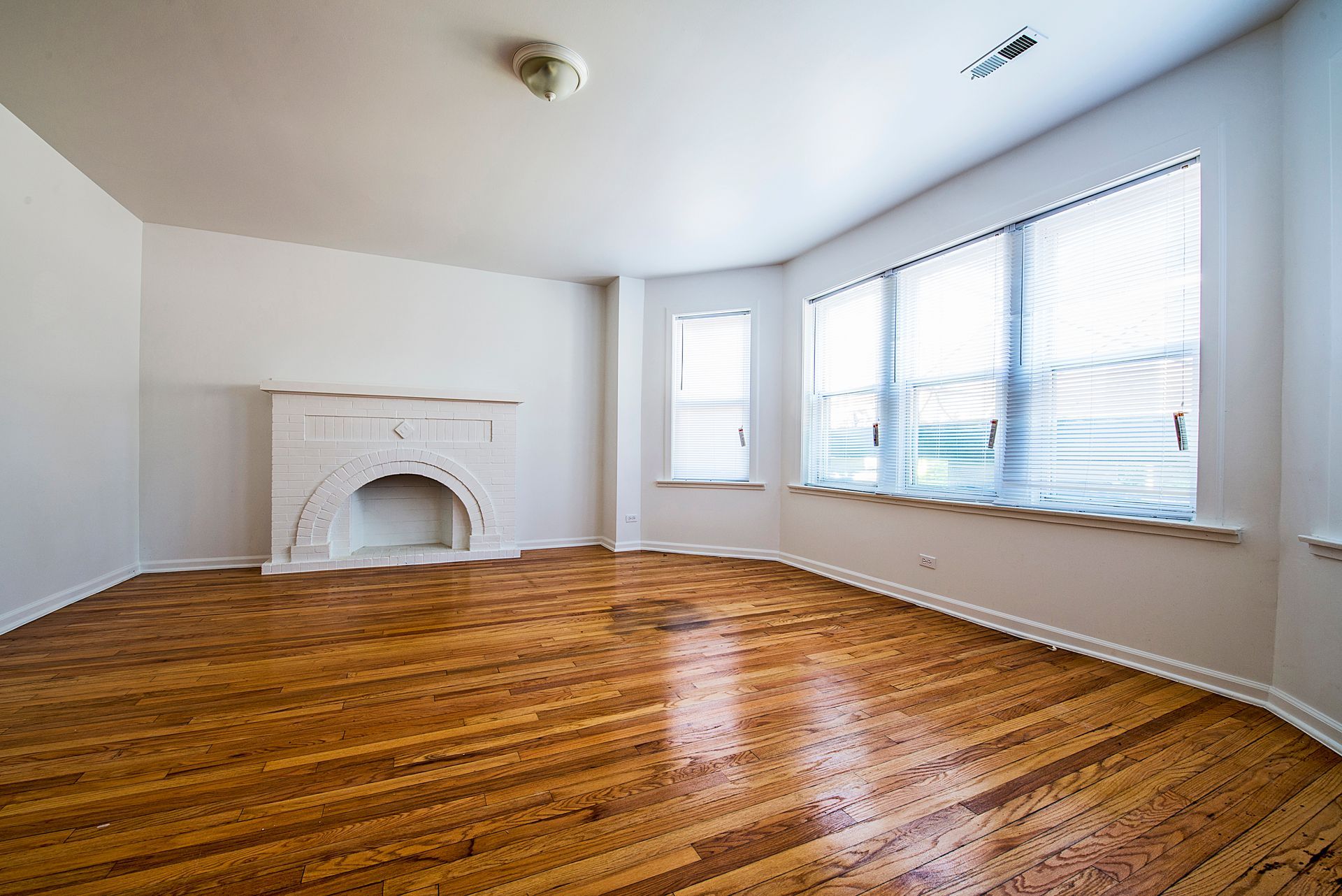 Empty room with hardwood floors, a white fireplace, and large windows with blinds.