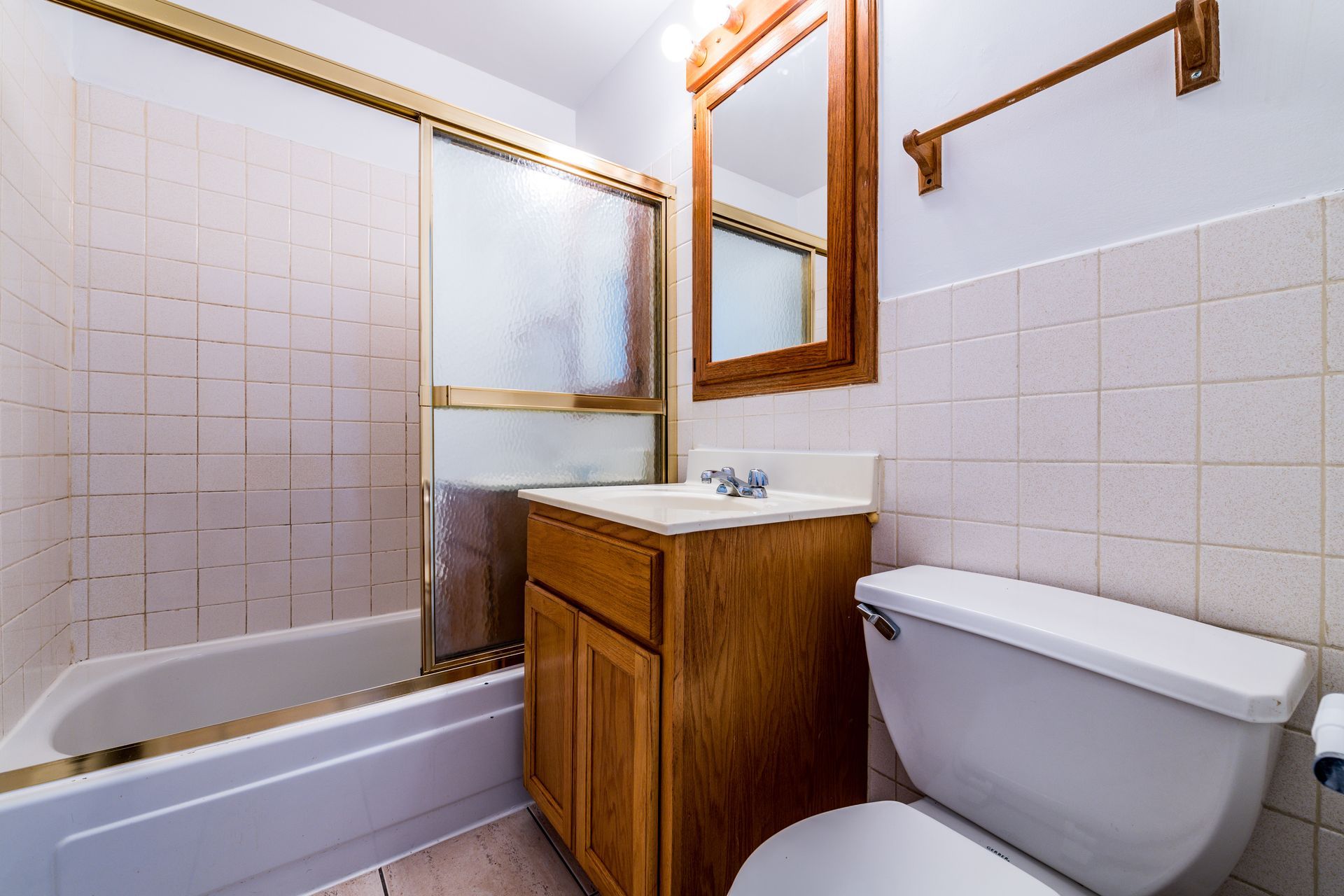 Small bathroom with white tile, a bathtub, wooden vanity and mirror, and a toilet.